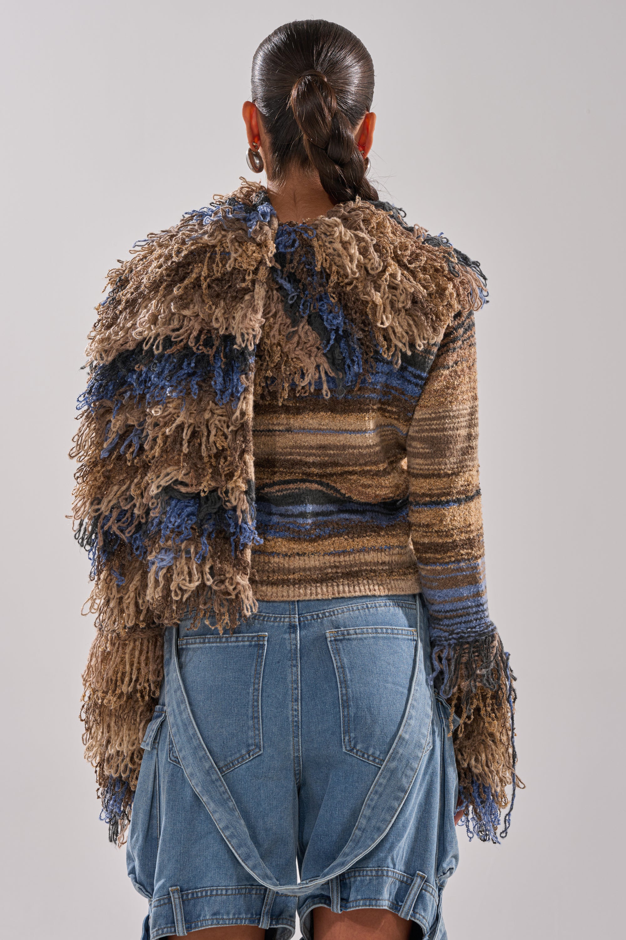 A person with long, dark braided hair is seen from behind wearing the ALWAYS ON MY MIND FRINGE SWEATER in brown and blue ombre tones with shaggy details, paired with high-waisted asymmetrical denim jeans with exposed pockets.