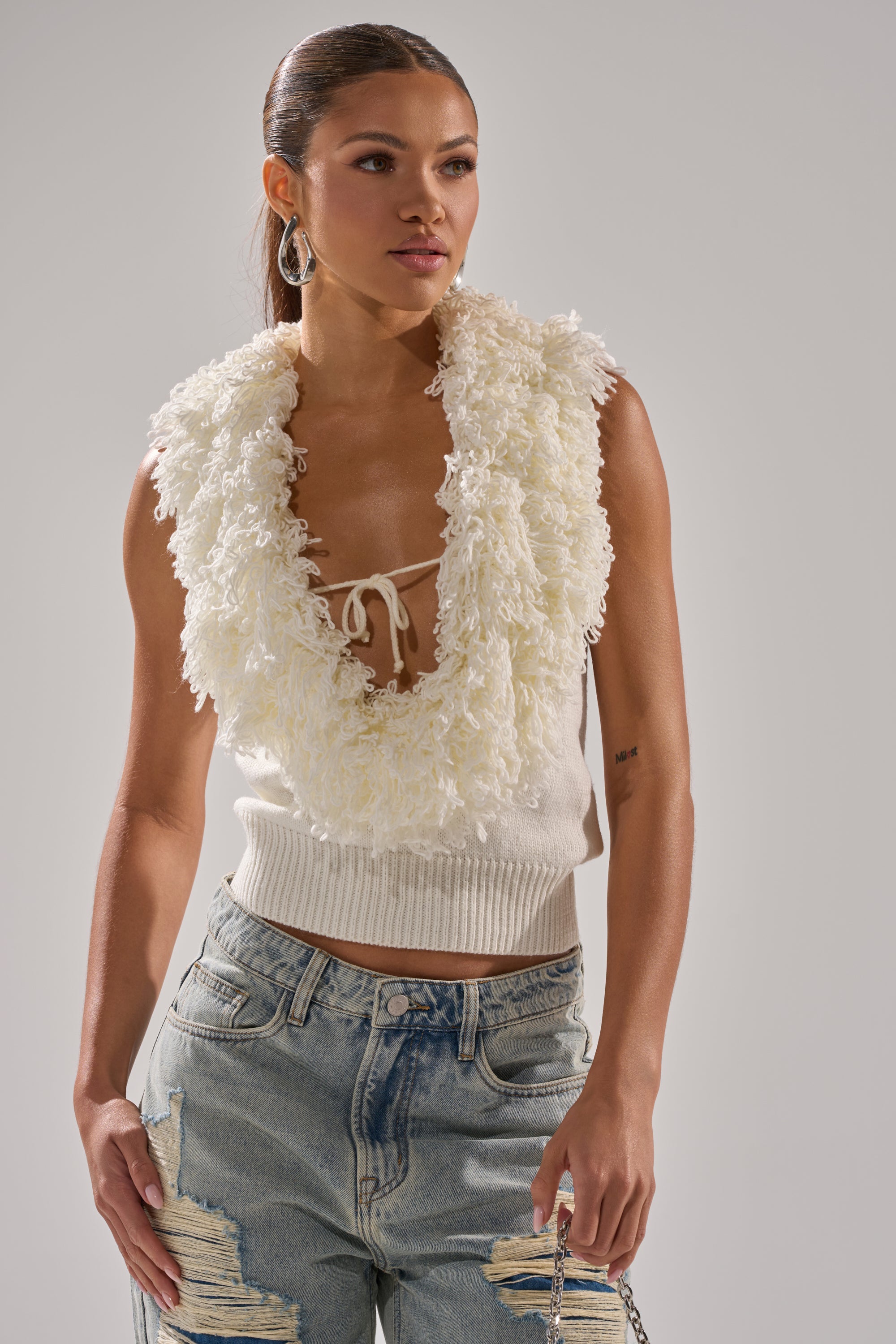 A woman wears the CARLY FRINGE OPEN FRONT SWEATER IN IVORY, featuring a fluffy fringe neckline, paired with distressed blue jeans, standing against a plain light background.