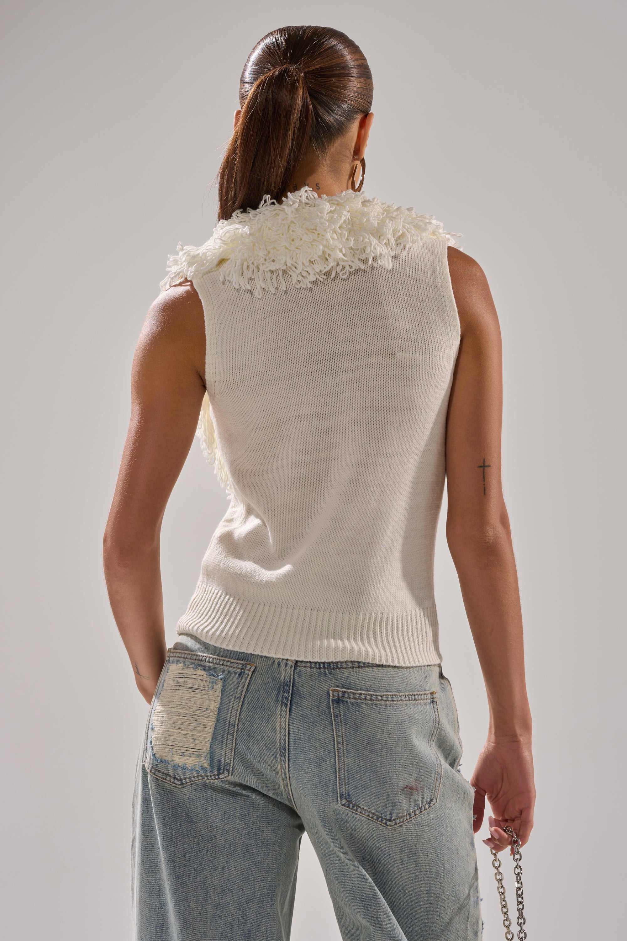 A woman with long brown hair in a ponytail wears the CARLY FRINGE OPEN FRONT SWEATER IN IVORY, featuring textured shoulder details, paired with light blue distressed jeans. She faces away and holds a chain strap bag.