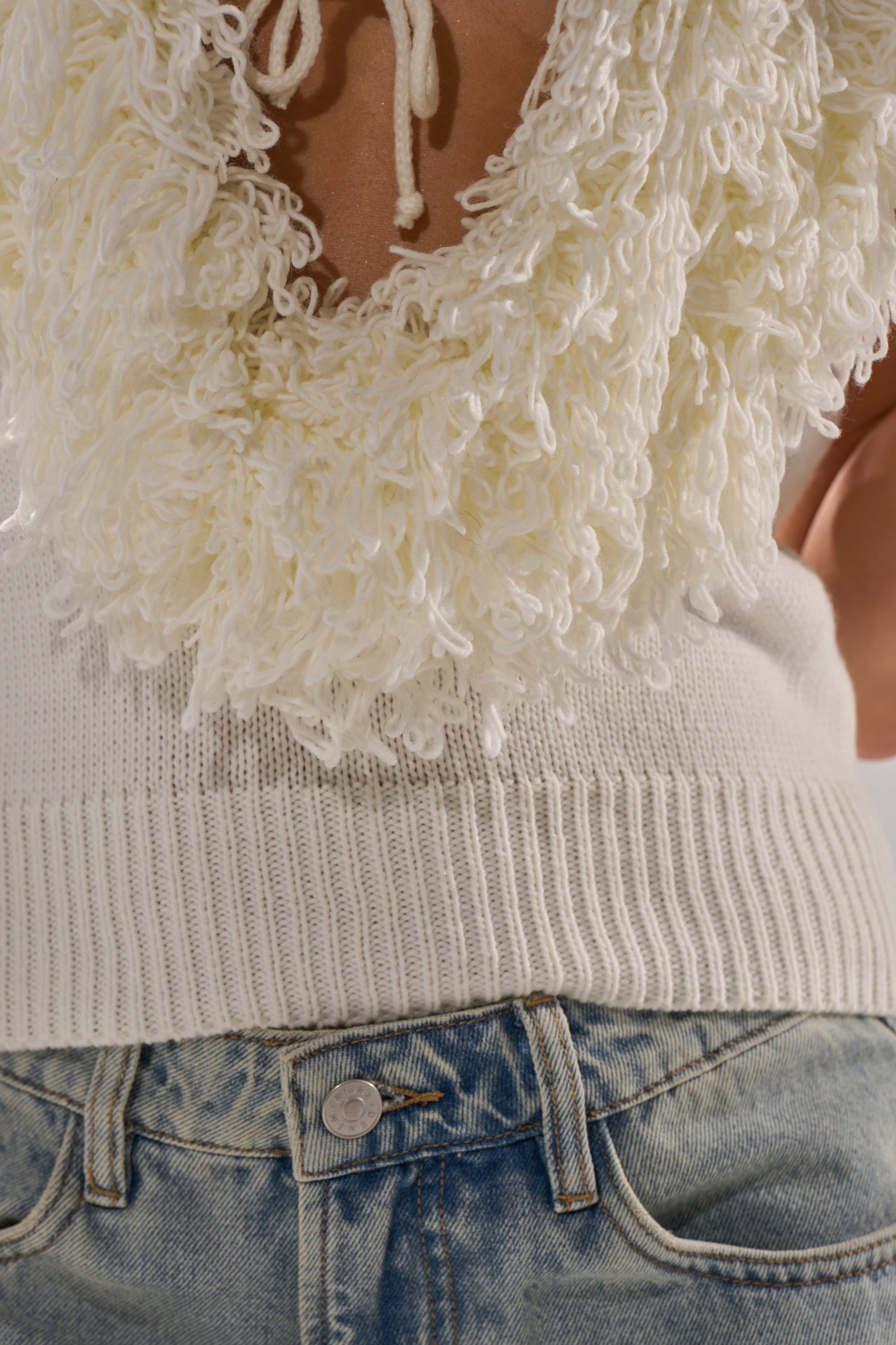 Close-up of a person wearing light blue denim jeans and the CARLY FRINGE OPEN FRONT SWEATER IN IVORY, featuring an AKIRA Label design with textured, looped yarn details and chic fringe around the neckline.