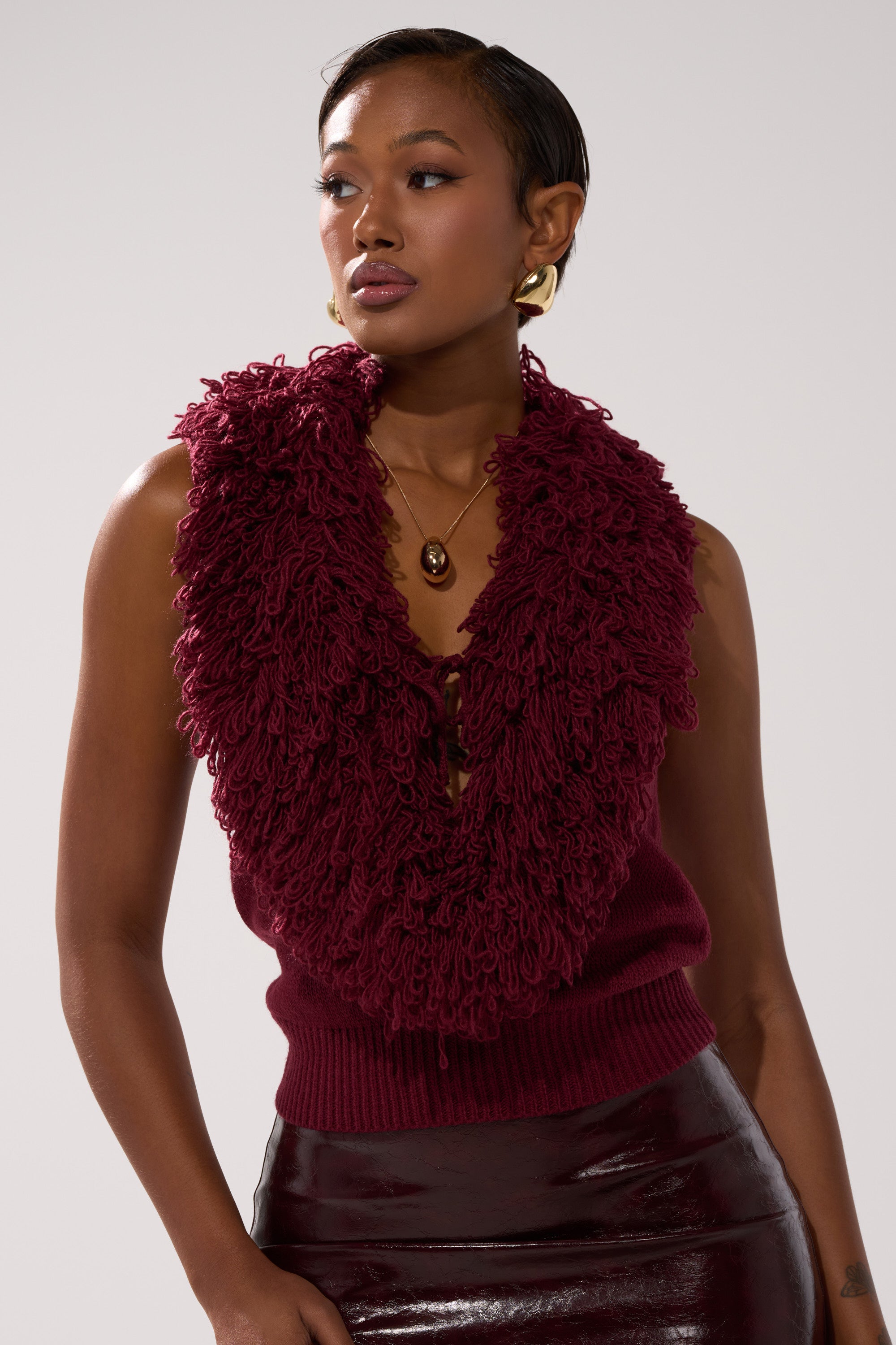A woman styles the CARLY FRINGE OPEN FRONT SWEATER, a burgundy sleeveless knit with fringe details, over a glossy dark burgundy skirt. She accessorizes with gold earrings, a pendant necklace, and sports short hair for an elegant look.