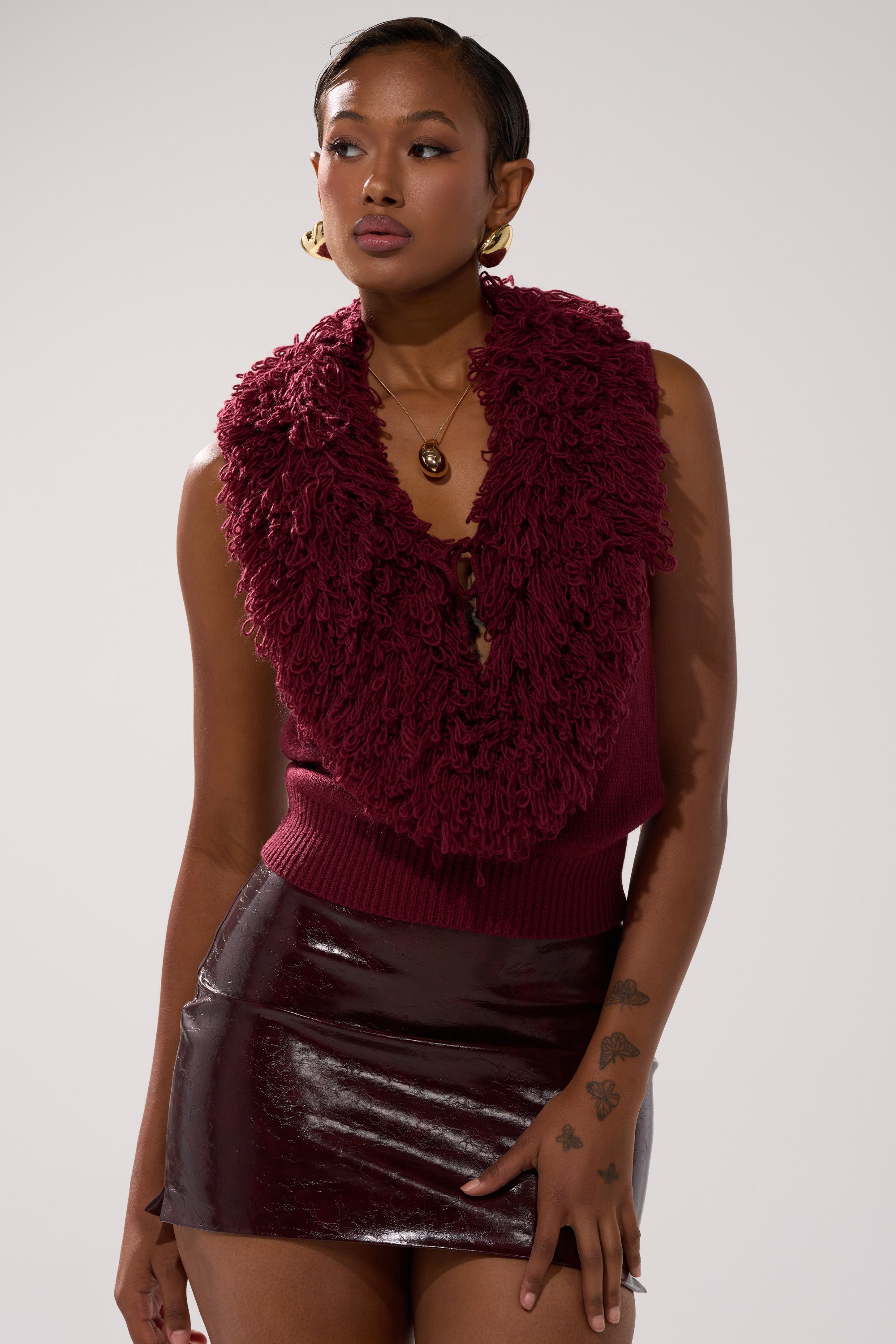 A woman wears a sleeveless burgundy top with a textured, shaggy collar inspired by the CARLY FRINGE OPEN FRONT SWEATER and pairs it with a shiny burgundy mini skirt, gold earrings, a pendant necklace, and shows tattoos on her left hand and arm.