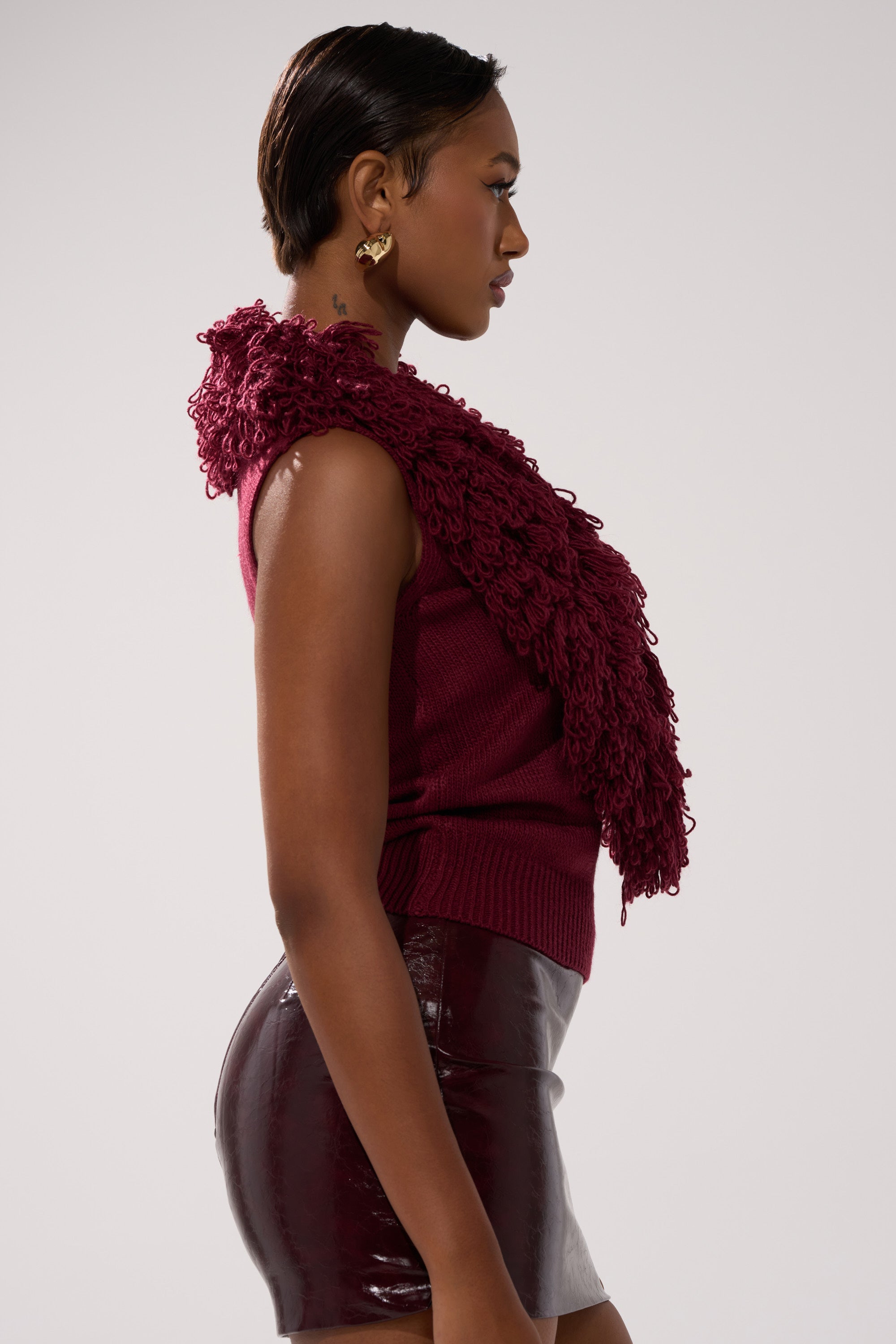 A woman in profile wears the CARLY FRINGE OPEN FRONT SWEATER, a sleeveless burgundy knit with textured fringe, paired with a shiny burgundy mini skirt. She has short hair and earrings, set against a light background.