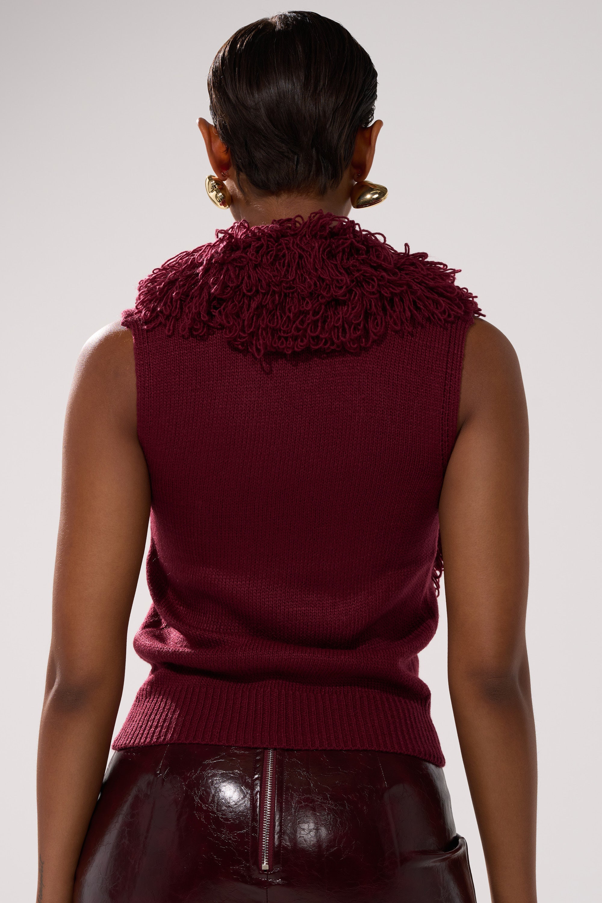 Someone wearing the CARLY FRINGE OPEN FRONT SWEATER, a sleeveless burgundy knit with textured detailing at the collar, paired with dark glossy pants, shown from behind against a plain background.