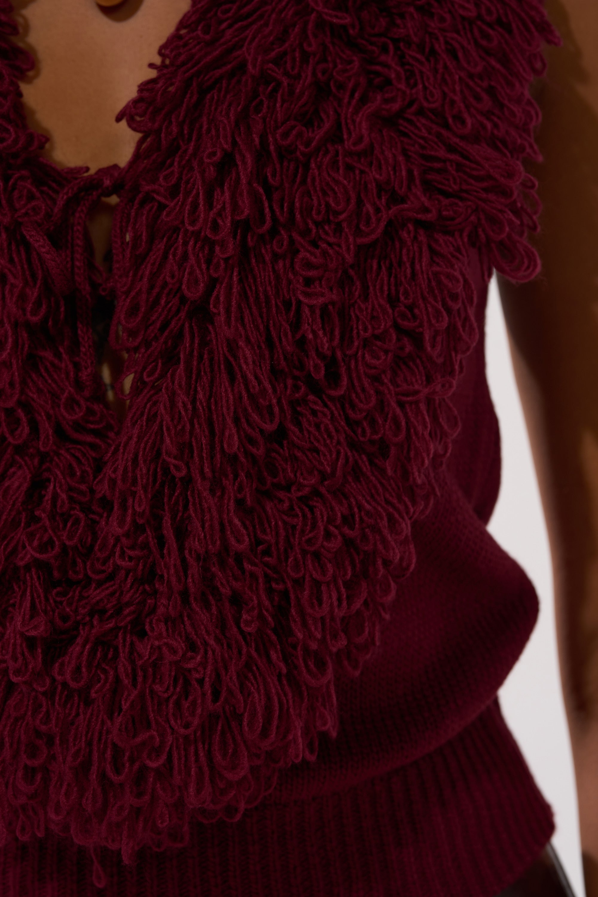 A close-up of a person wearing the CARLY FRINGE OPEN FRONT SWEATER in burgundy, featuring thick, textured looped yarn fringe around the neckline and chest area.