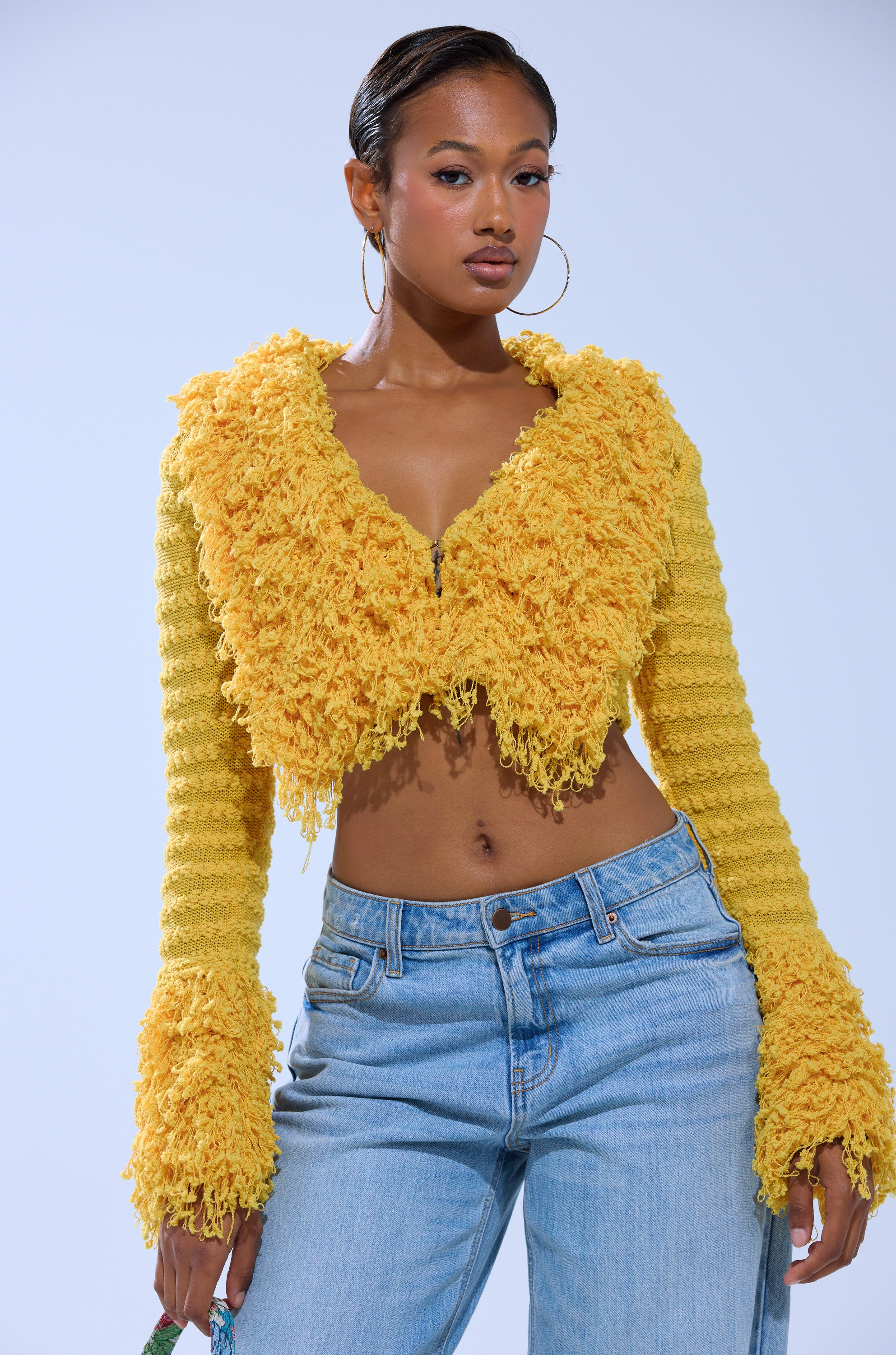 A woman poses confidently against a plain light background in the SUNSHINE QUEEN FRINGE SWEATER, a bright yellow shaggy knit cropped top with flared sleeves, paired with light blue high-waisted jeans and hoop earrings, her hair styled back. 0