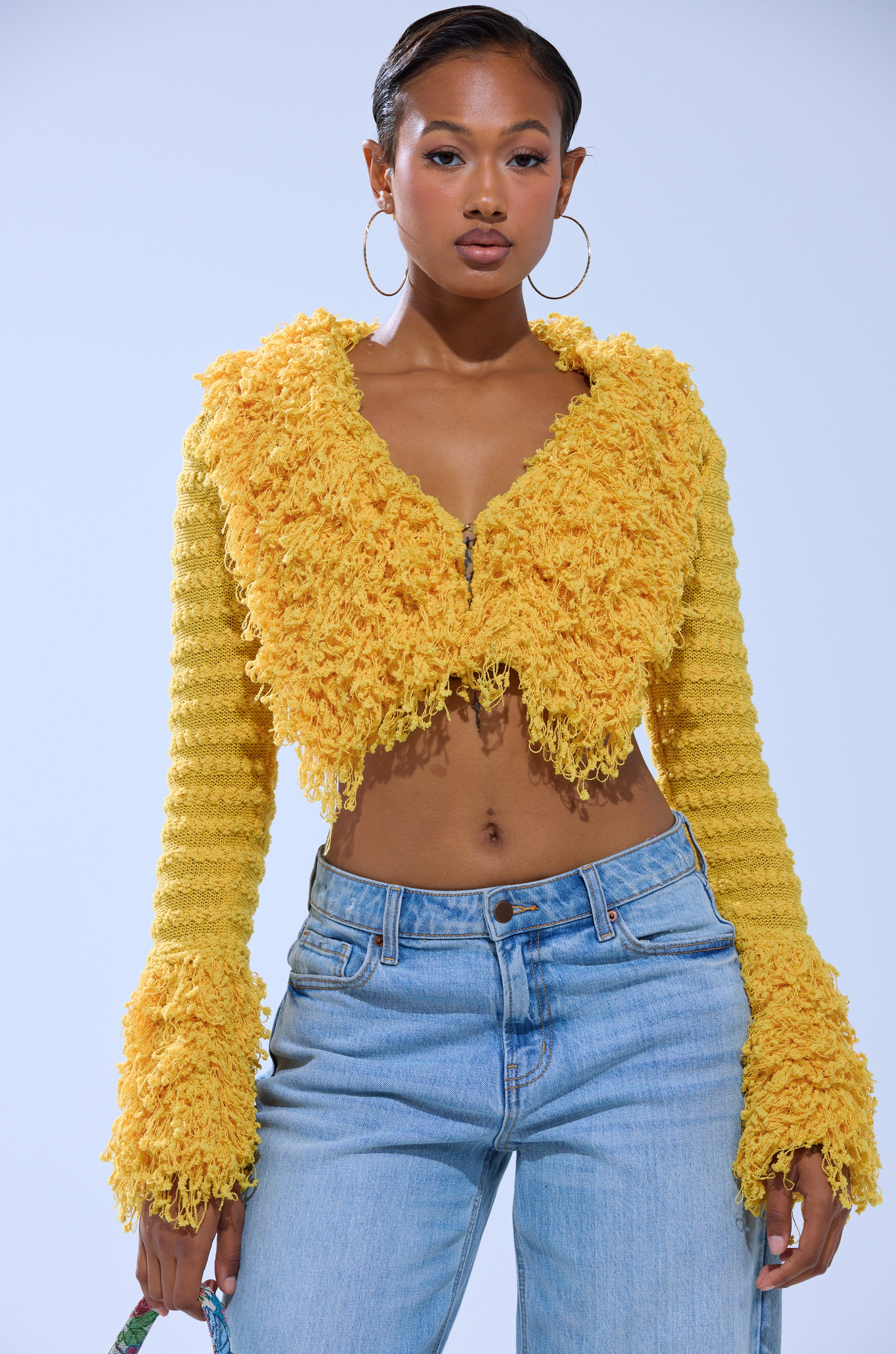 A woman stands confidently against a plain background, wearing the SUNSHINE QUEEN FRINGE SWEATER—a yellow textured knit cropped cardigan with flared sleeves—paired with light blue jeans. 1