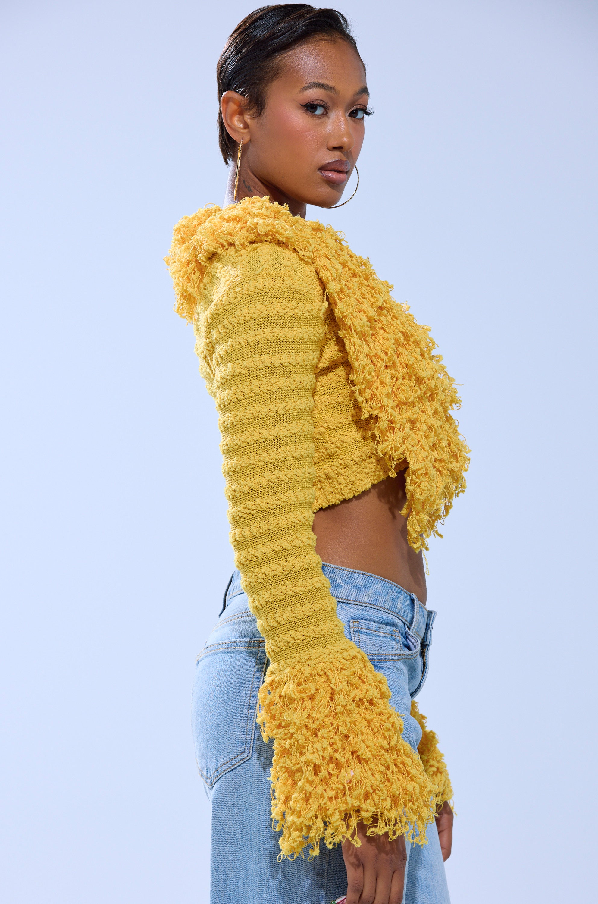 A woman in profile wears the SUNSHINE QUEEN FRINGE SWEATER, a textured knit yellow cropped top with flared sleeves, paired with light blue jeans. She accessorizes with hoop earrings and has slicked-back hair against a plain light background. 2