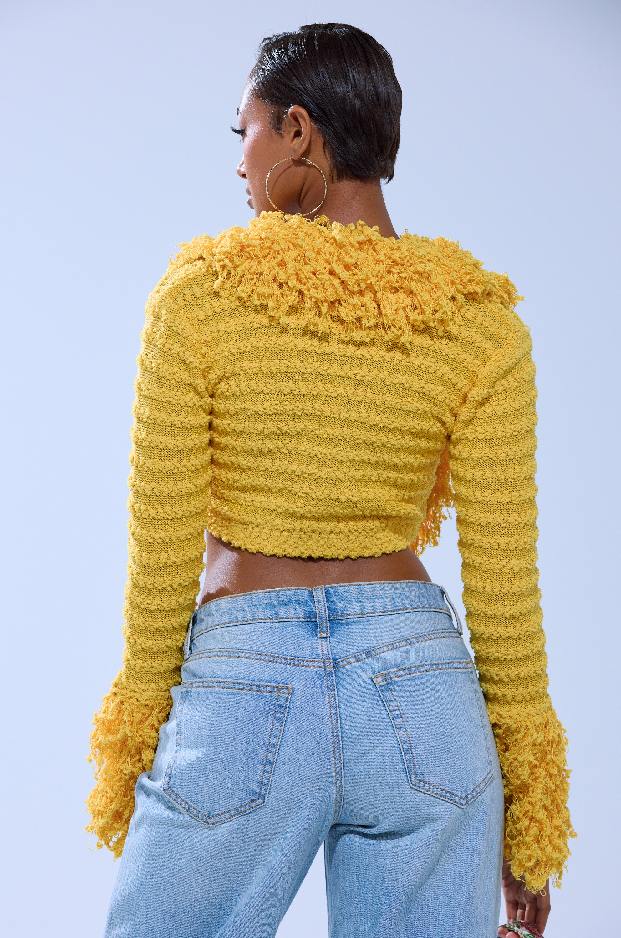 A person with short dark hair is seen from the back, wearing the SUNSHINE QUEEN FRINGE SWEATER in yellow and light blue high-waisted jeans, standing against a plain light background. 3