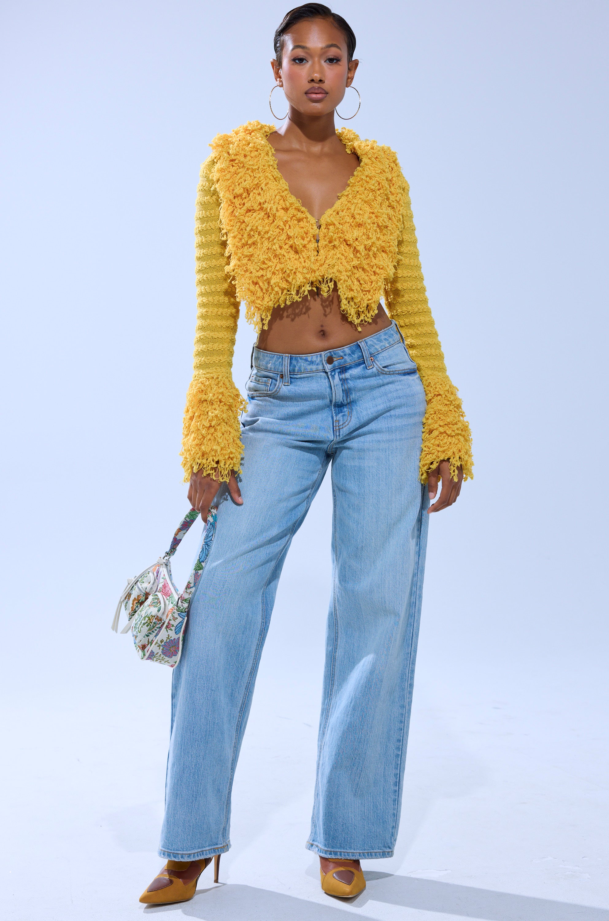 A woman stands confidently in the SUNSHINE QUEEN FRINGE SWEATER, paired with light blue wide-leg jeans, yellow heels, large hoop earrings, and a small floral handbag against a light background. 4