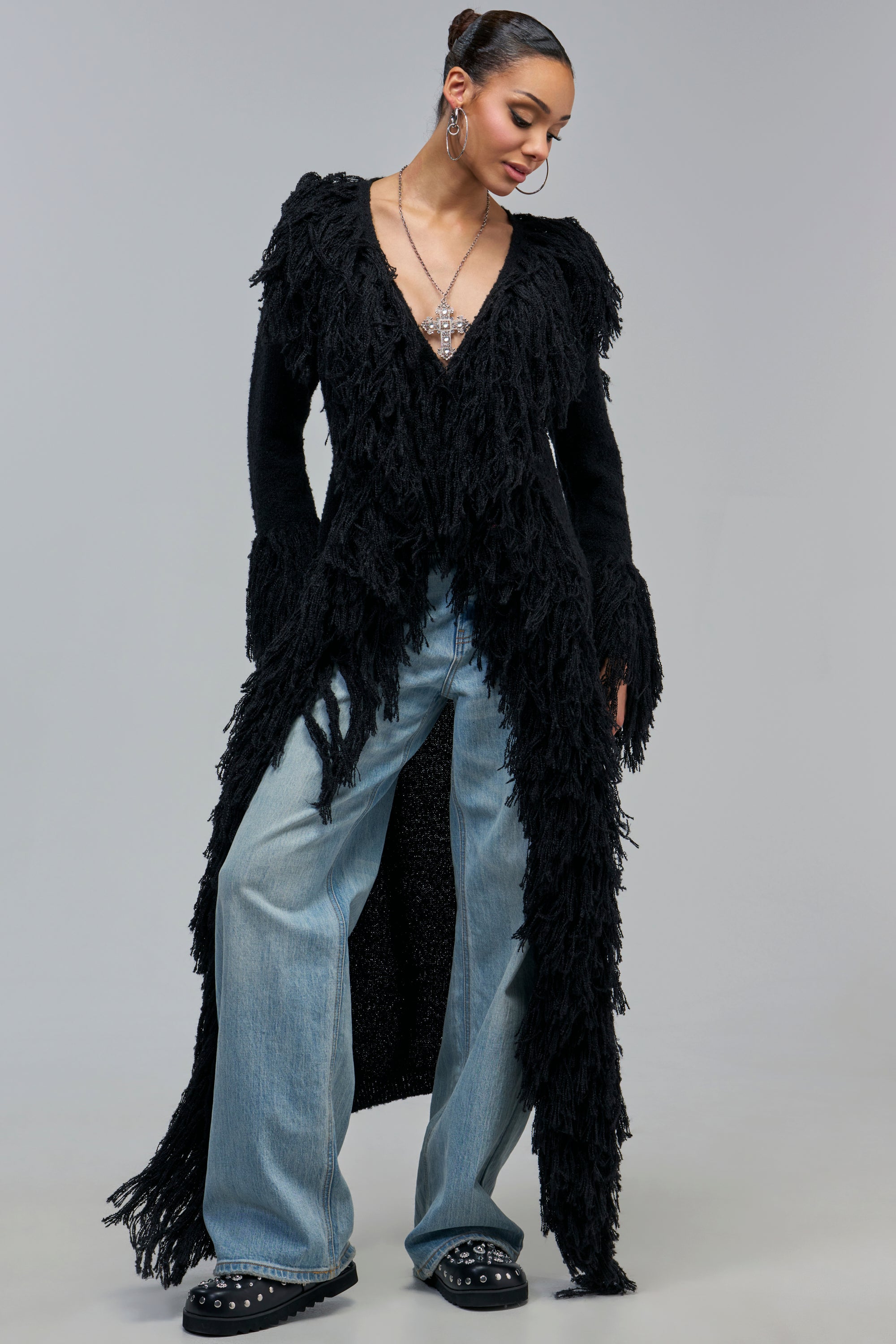 A person wearing a CANT GET ENOUGH FRINGE CARDIGAN IN BLACK, light-wash blue jeans, and black studded shoes.