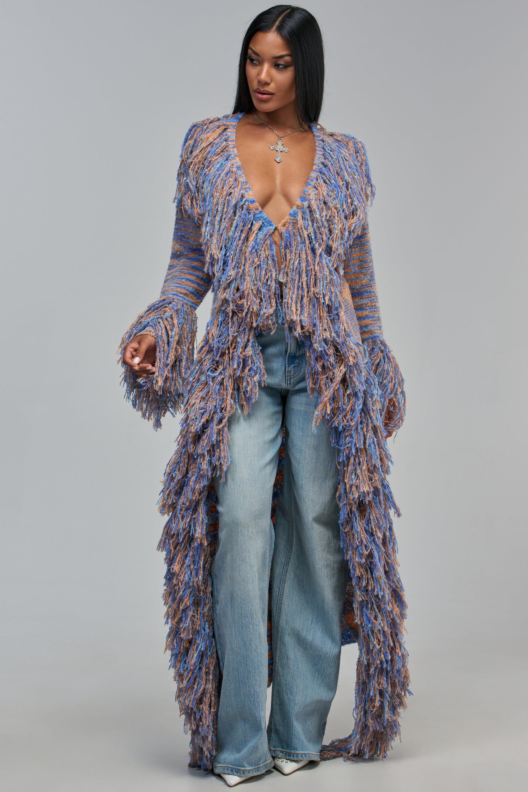 A person wears the CANT GET ENOUGH FRINGE CARDIGAN IN BLUE MULTI, an open, longline blue and orange knit cardigan covered in fringe, paired with light wash blue jeans for a stylish fall layering look.