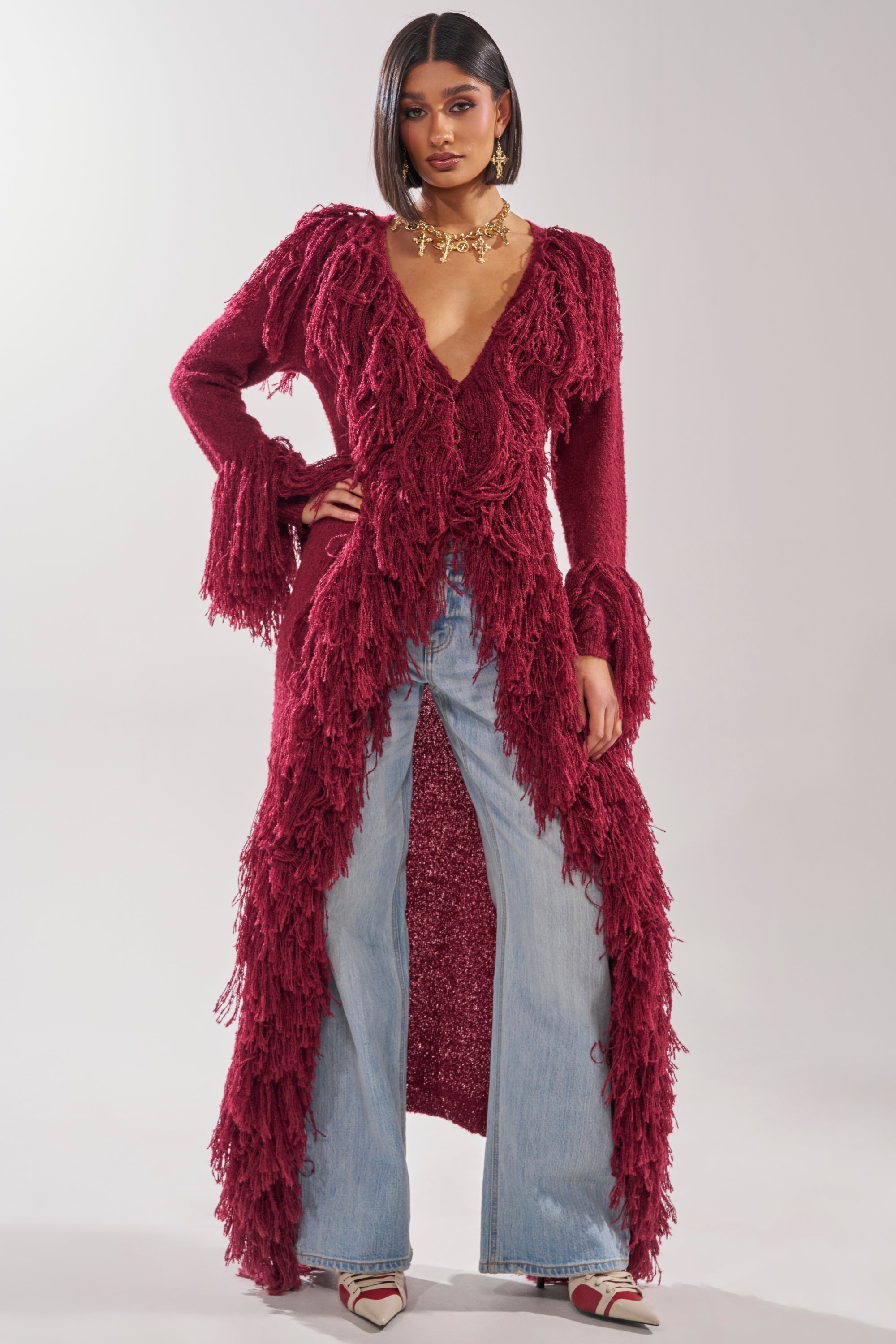 A woman exudes confidence in the CAN'T GET ENOUGH FRINGE CARDIGAN, styled with a plunging neckline, light-wash flared jeans, pointed heels, and a gold statement necklace against a plain background. 0