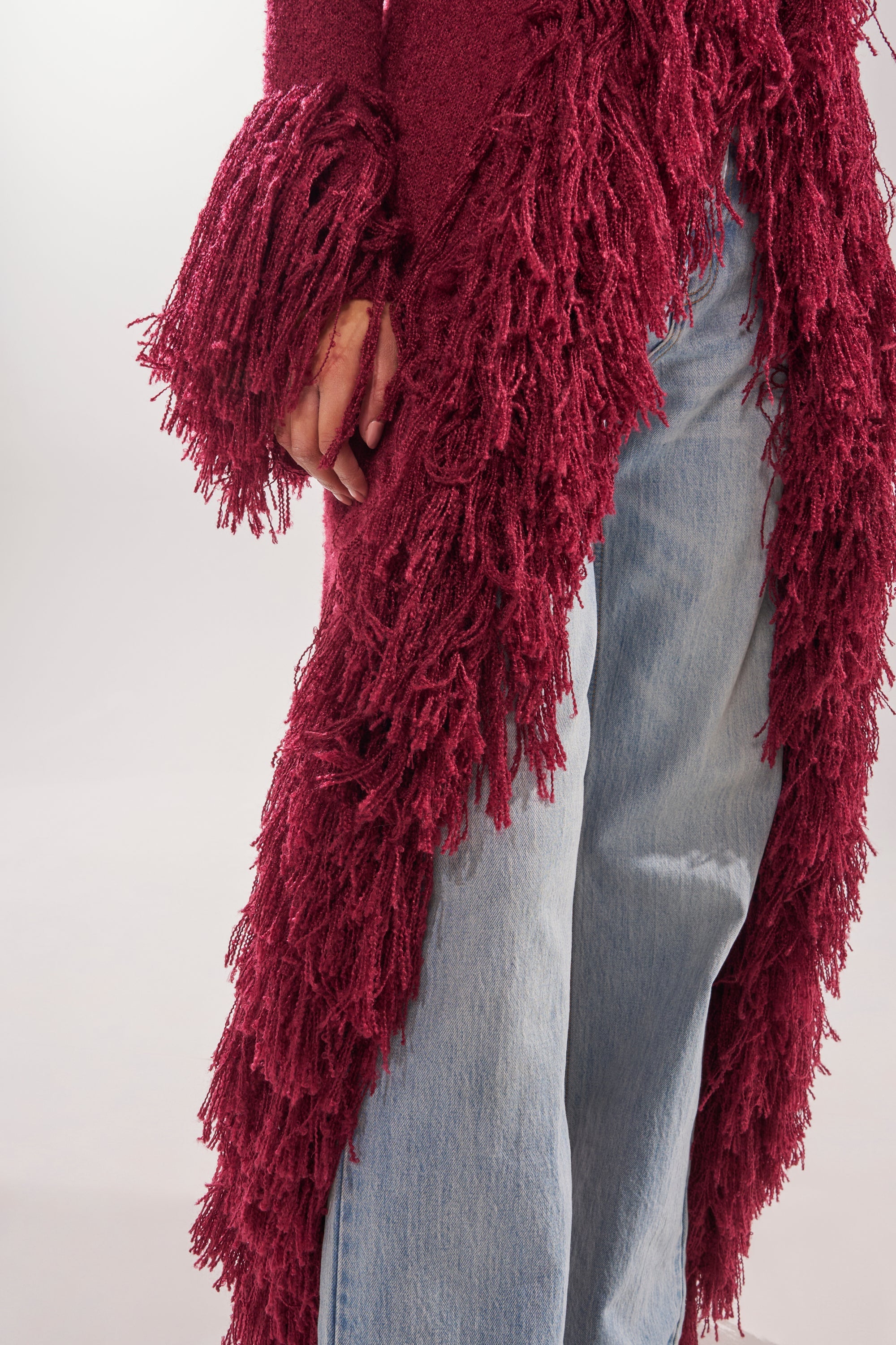 A person wears the CAN'T GET ENOUGH FRINGE CARDIGAN—burgundy, longline, with thick fringe on sleeve and side—paired with light blue jeans, photographed from the shoulders down against a plain background. 9