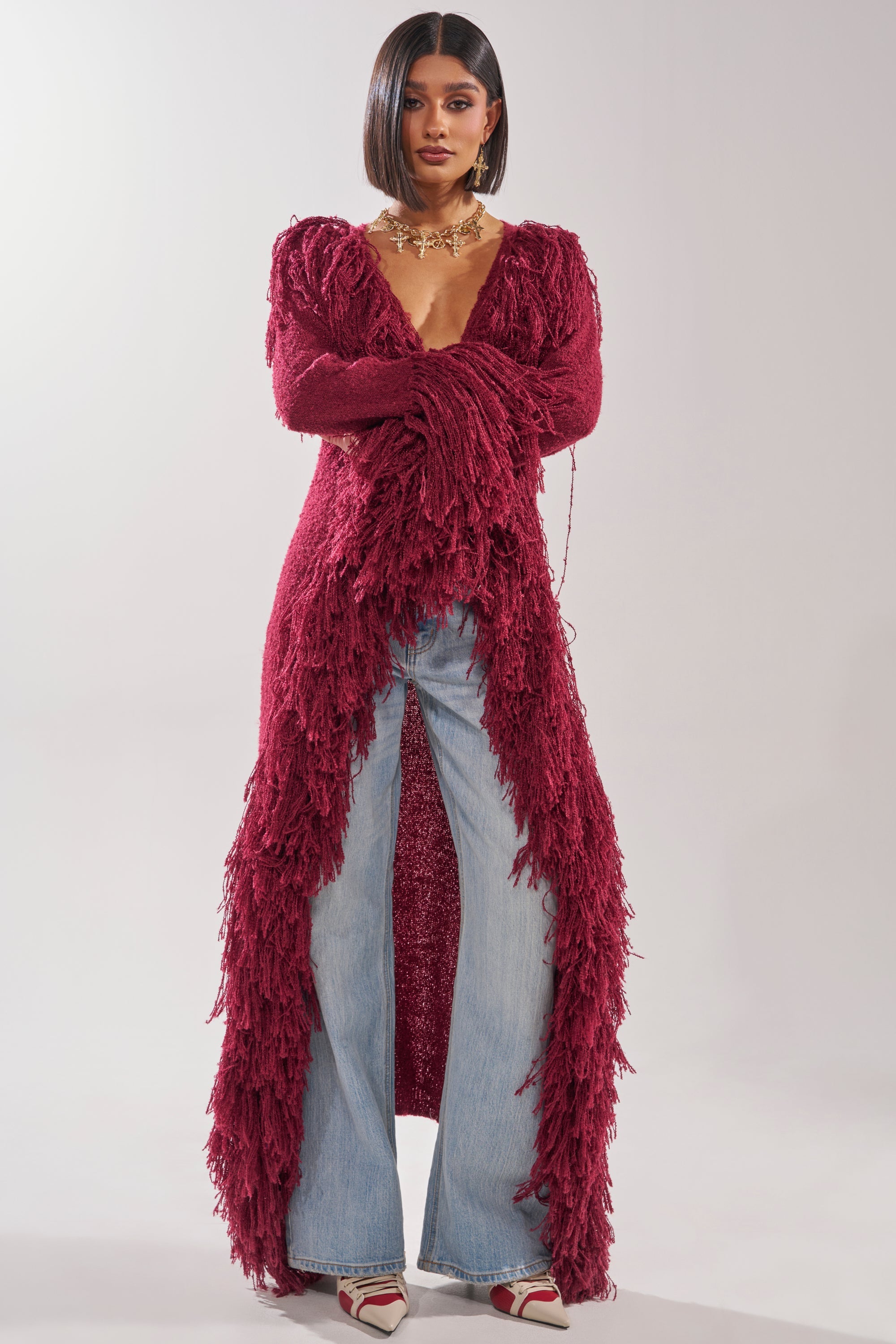 A woman stands facing forward in the CAN'T GET ENOUGH FRINGE CARDIGAN, a burgundy longline style with fringe details, paired with light blue wide-leg jeans, pointed-toe shoes, and a chunky gold necklace. 1