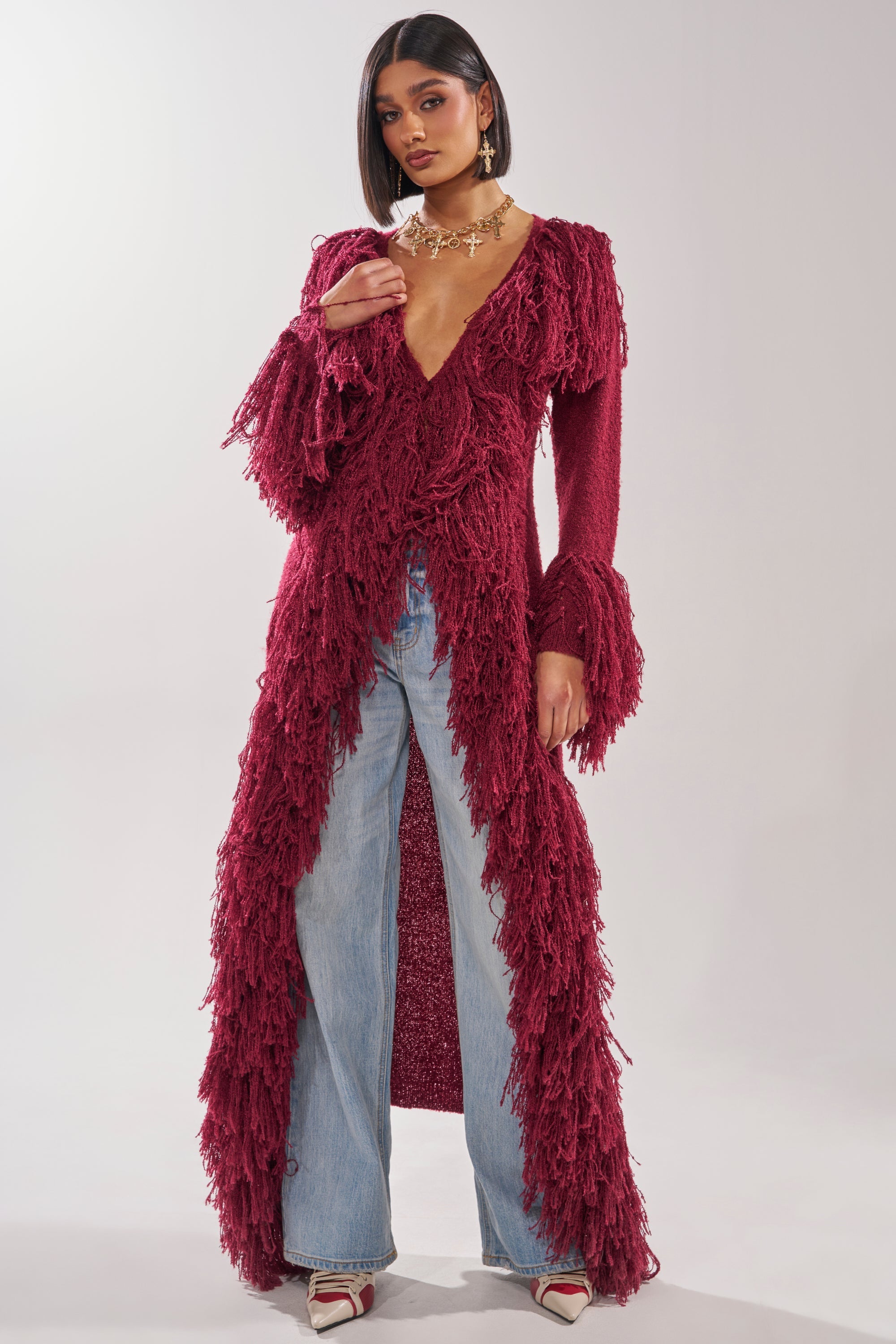 A woman wears the CAN'T GET ENOUGH FRINGE CARDIGAN in burgundy as a longline layer over light blue wide-leg jeans and white pointed-toe heels, standing against a plain light background. 3