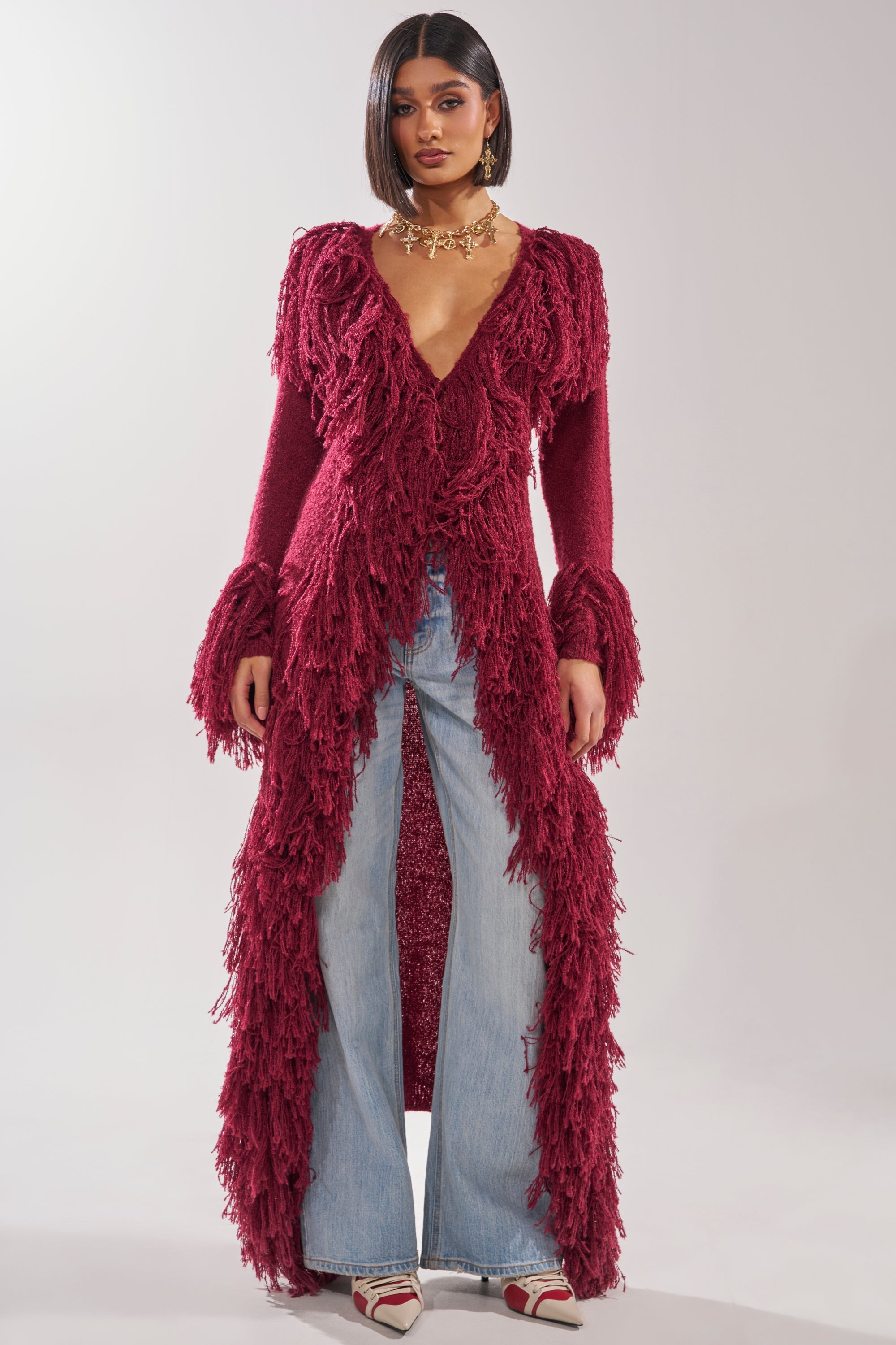 A woman stands against a light background wearing the CAN'T GET ENOUGH FRINGE CARDIGAN, styled with light blue wide-leg jeans, pointed-toe shoes, and a chunky gold necklace. 4
