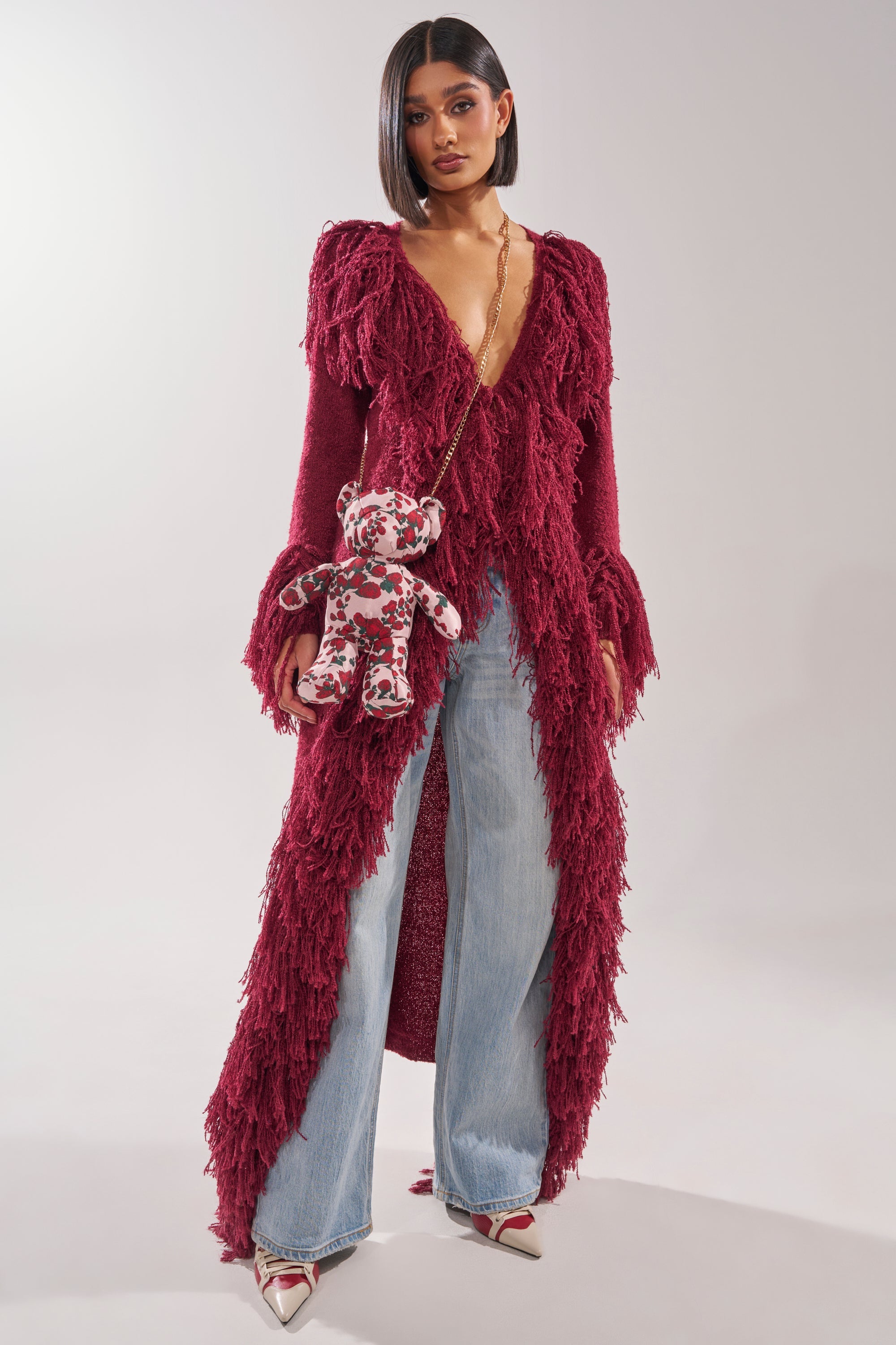 A woman wears the CAN'T GET ENOUGH FRINGE CARDIGAN in burgundy over light blue wide-leg jeans and pointed heels, carrying a teddy bear-shaped crossbody bag with a pink and red floral pattern. 5
