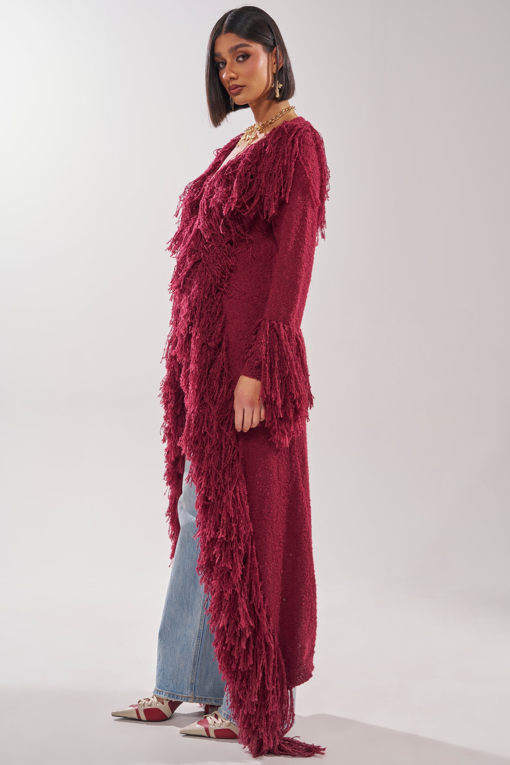 A woman with a bob haircut models the CAN'T GET ENOUGH FRINGE CARDIGAN in magenta, paired with blue jeans, white sneakers, and gold jewelry, standing against a light gray background. 6