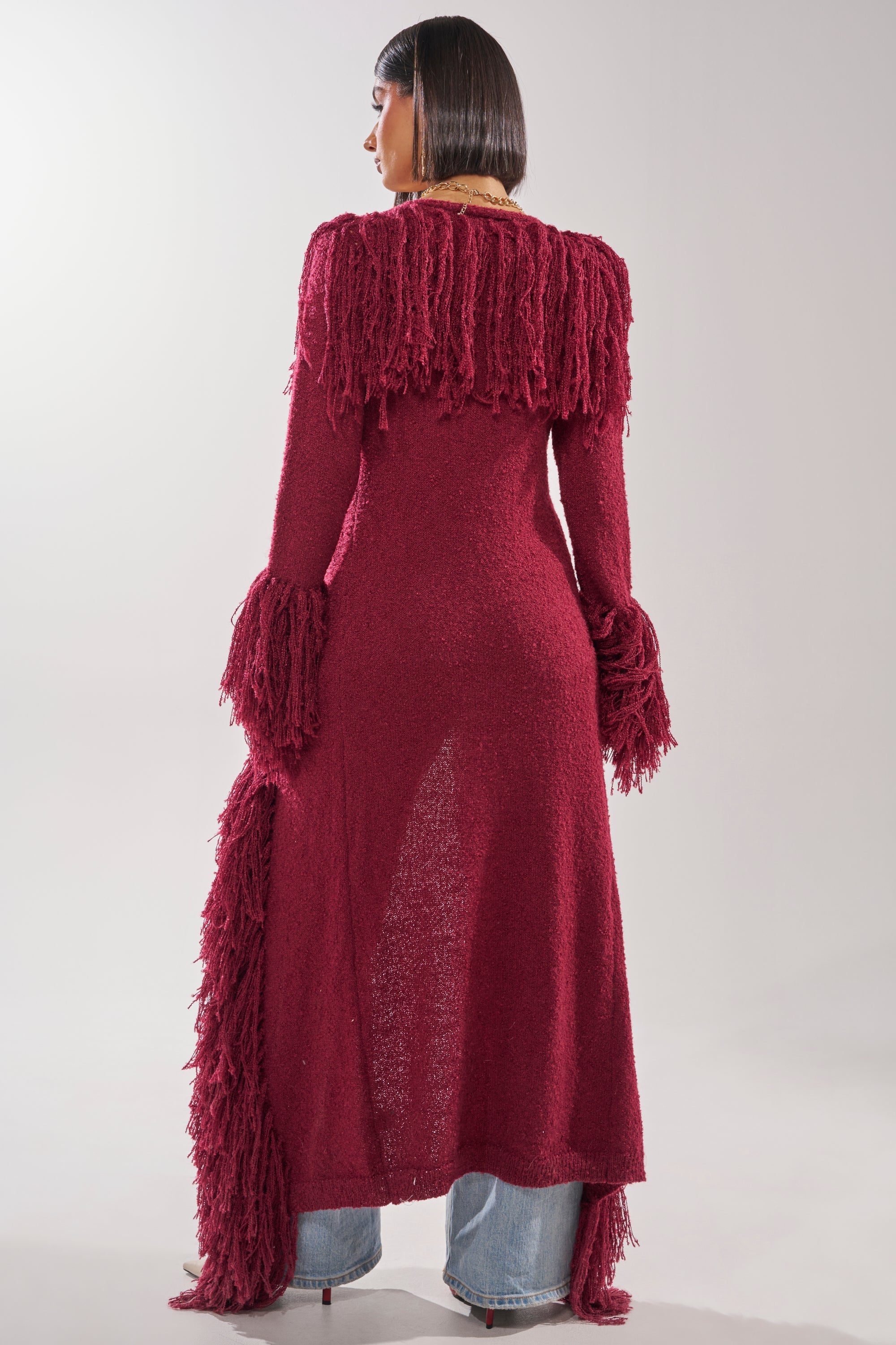 A woman with a sleek bob and blue jeans stands facing away, wearing the CAN'T GET ENOUGH FRINGE CARDIGAN in burgundy, featuring fringe details on the shoulders, sleeves, and one side. The background is plain and light. 7
