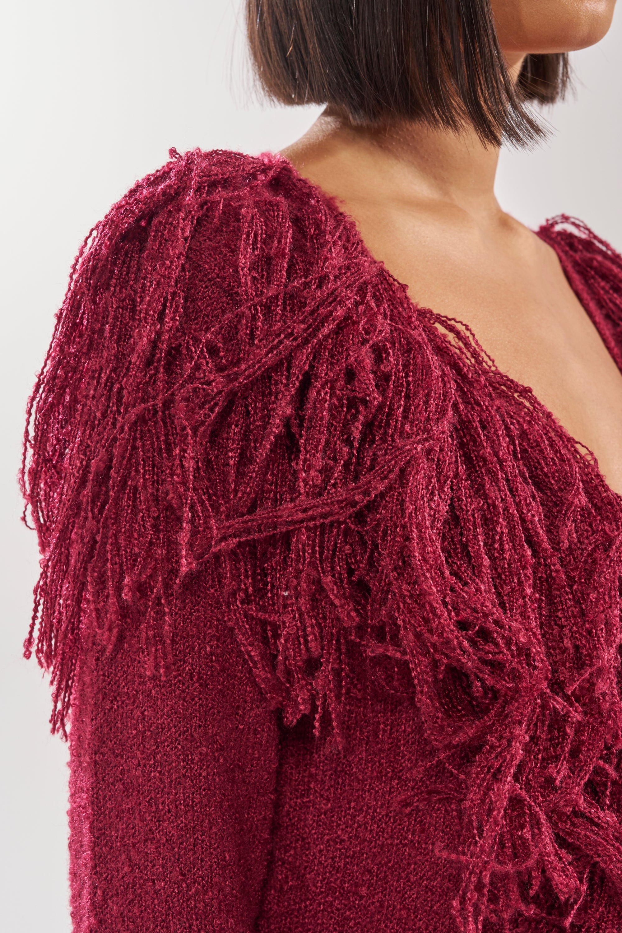 A woman with a short bob showcases the CAN'T GET ENOUGH FRINGE CARDIGAN in textured deep red with thick fringe on the shoulder and upper sleeve; only her shoulder, neck, and part of her face are visible. 8