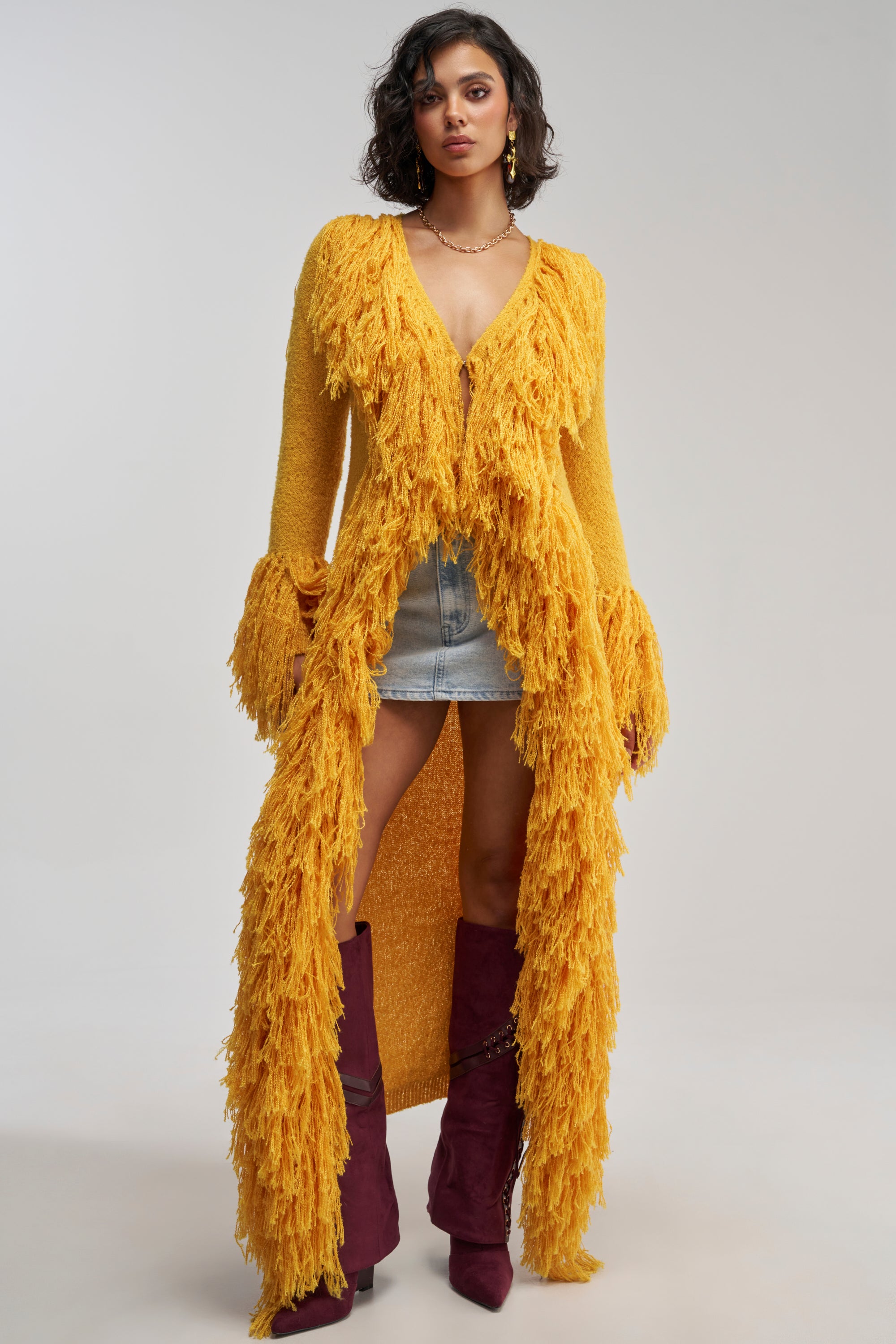 Wearing the VICE CITY FRINGE CARDIGAN in mustard, a white top, denim shorts, and tall burgundy boots, a person stands against a plain background.