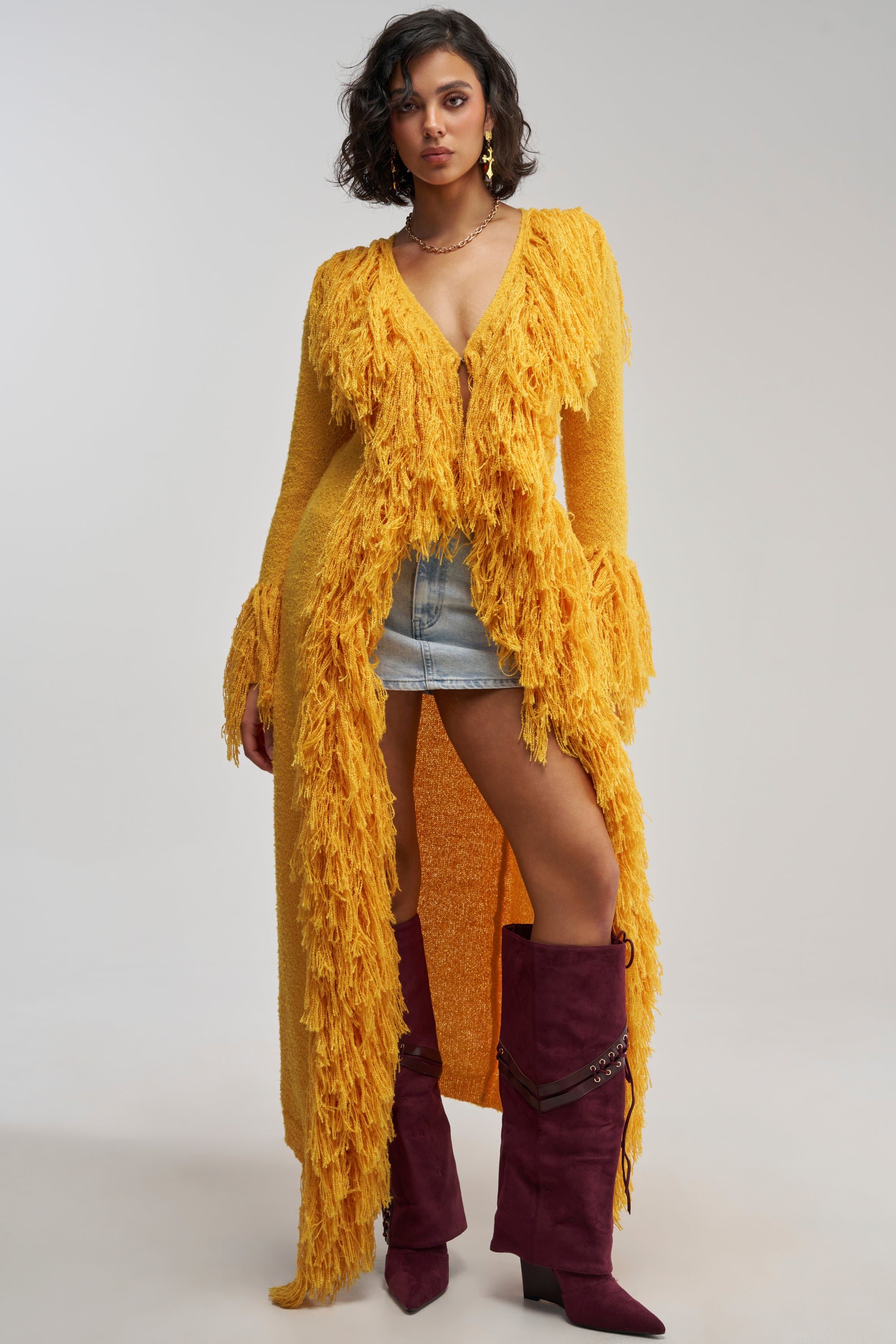 Wearing the VICE CITY FRINGE CARDIGAN in a vibrant yellow duster length, a woman stands confidently against a plain background, pairing the statement cardigan with a low-cut top, denim shorts, and tall maroon boots.