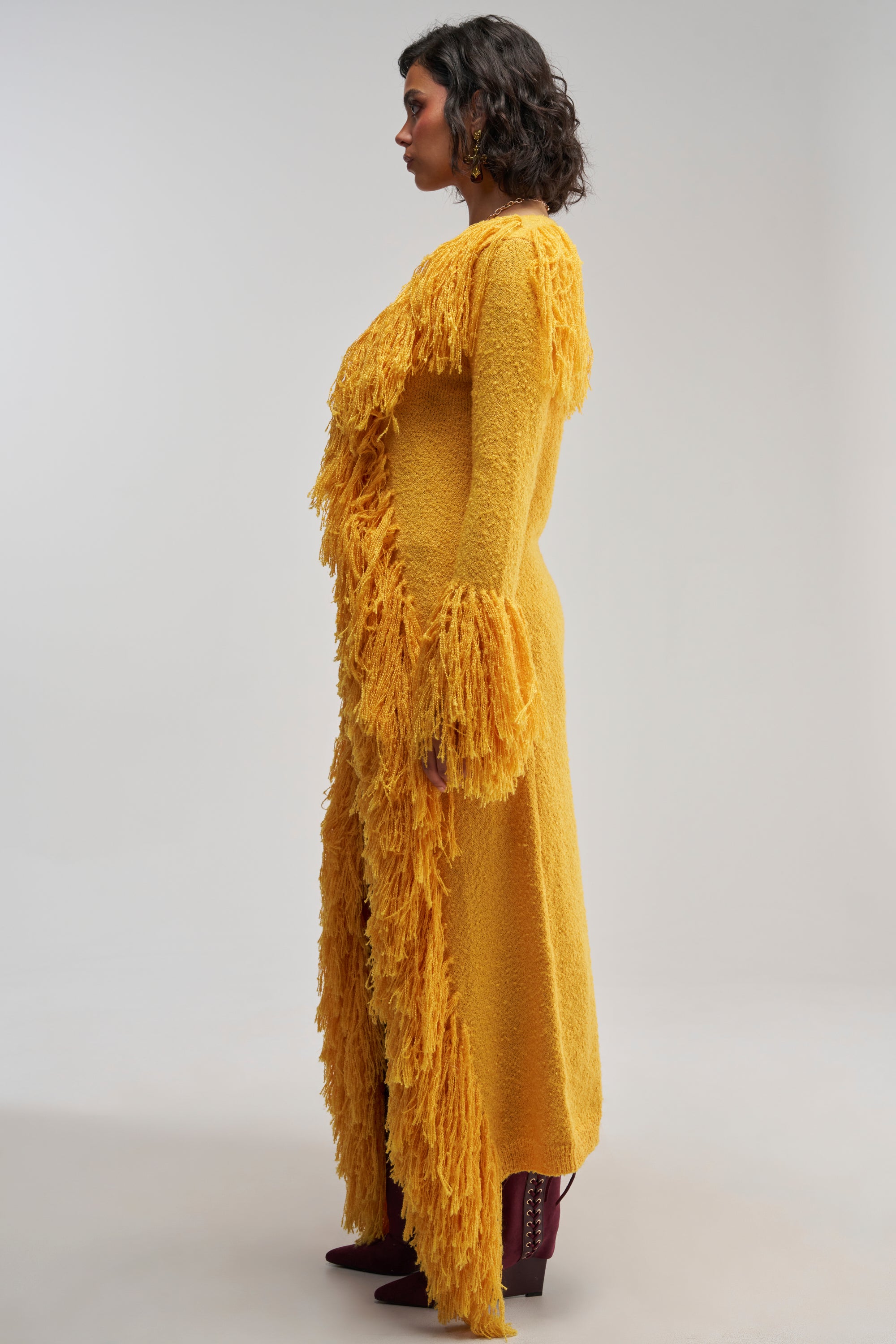 A person stands in profile wearing a long, textured yellow dress with fringe details and flared sleeves, styled with the VICE CITY FRINGE CARDIGAN and burgundy boots, against a plain light background.