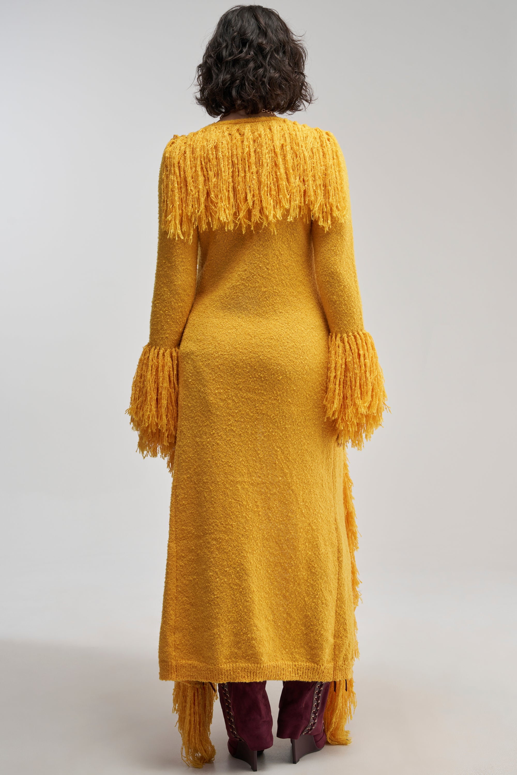 A person stands facing away, wearing the VICE CITY FRINGE CARDIGAN—a long yellow textured piece with fringed details on the shoulders and sleeves—styled with maroon boots. The look, reminiscent of a duster-length cardigan, is set against a plain, light background.