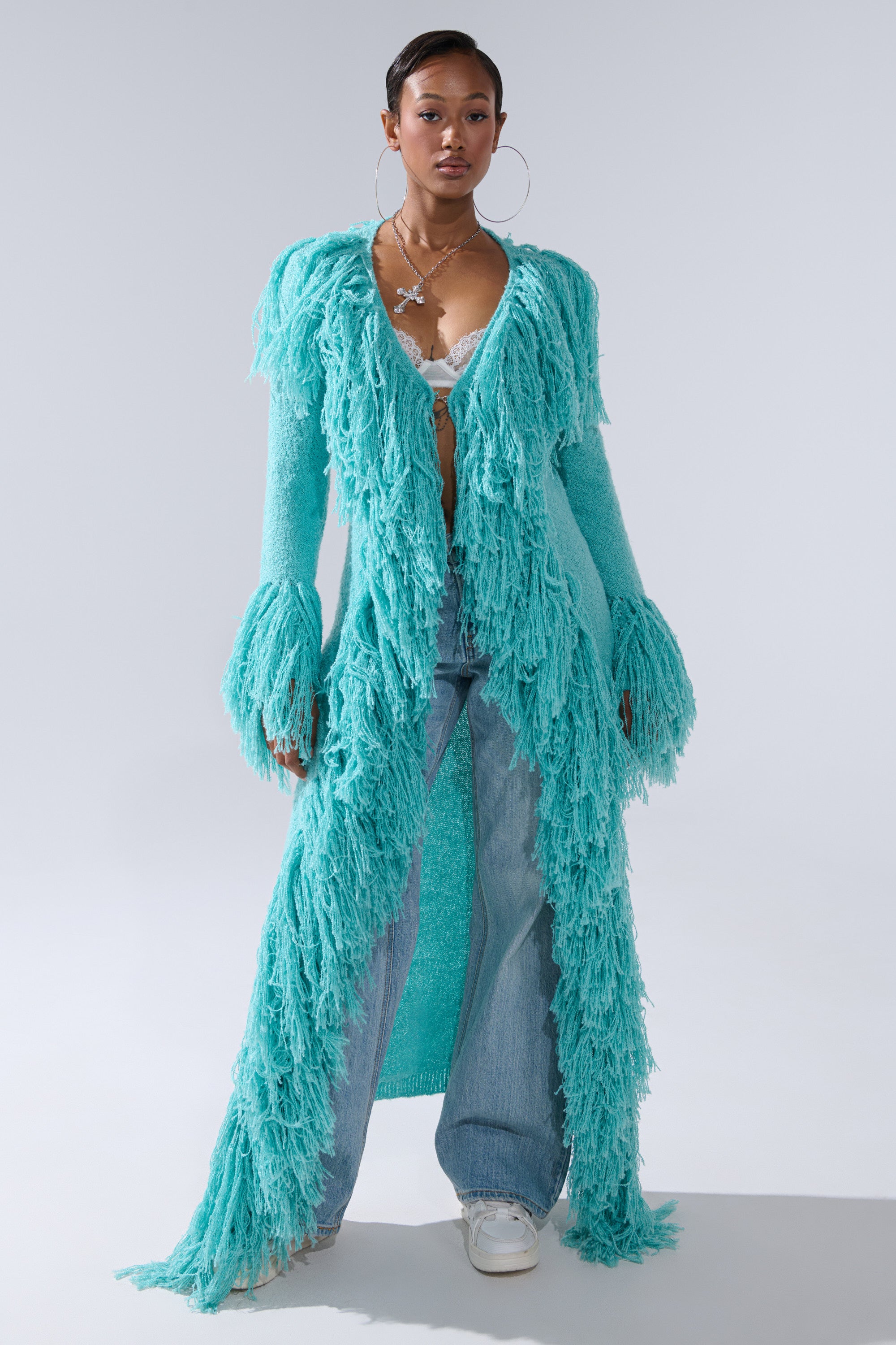 A woman models the BURRR IT'S COLD OUTSIDE FRINGE OVERSIZED SWEATER IN TURQUOISE over a white lace bralette, paired with wide-leg blue jeans and white sneakers, accessorized with large hoop earrings and a statement necklace.