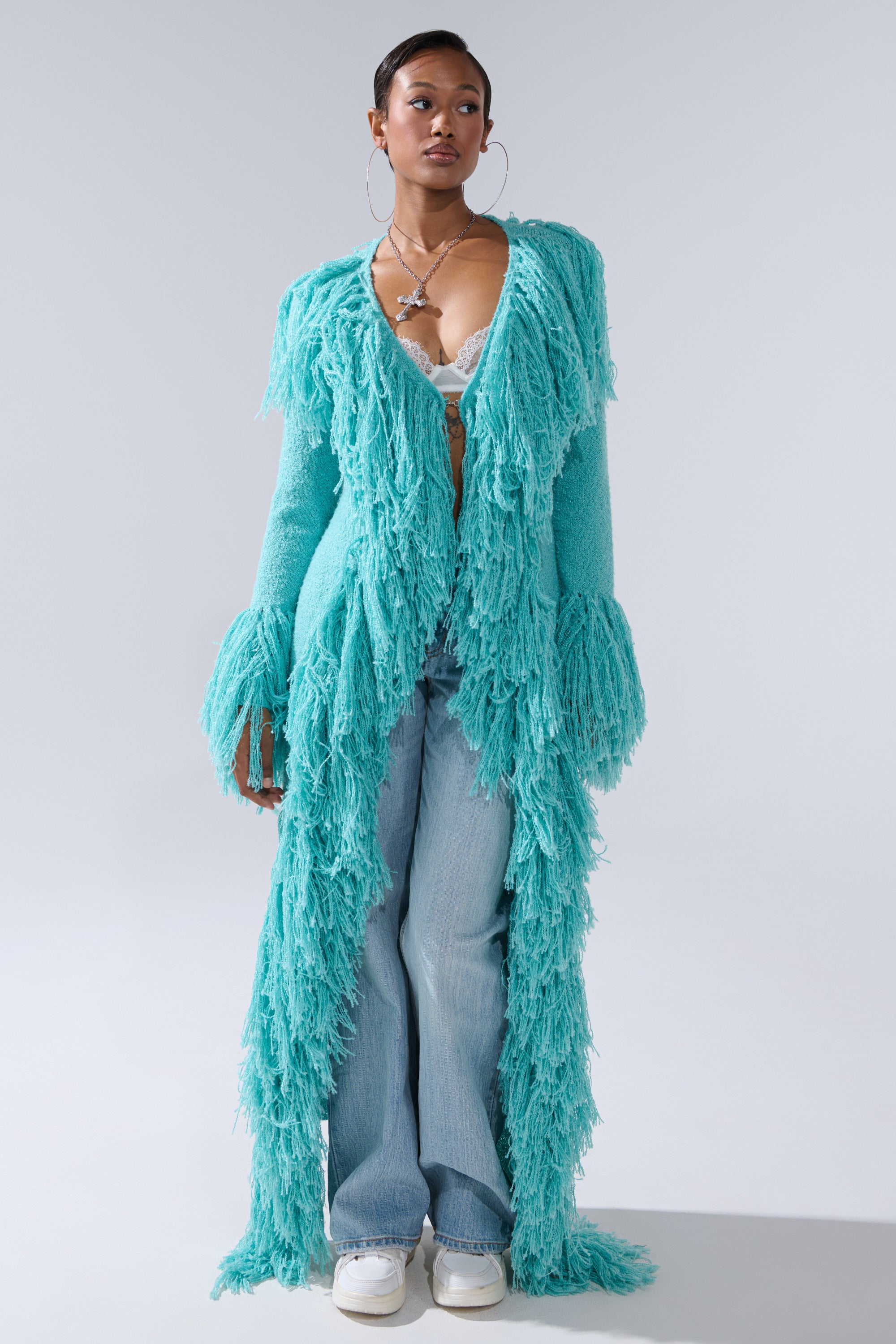 A woman wears the BURRR IT'S COLD OUTSIDE FRINGE OVERSIZED SWEATER IN TURQUOISE over a lacy bralette and wide-leg light blue jeans, styled with white sneakers and large hoop earrings, standing against a plain light background.