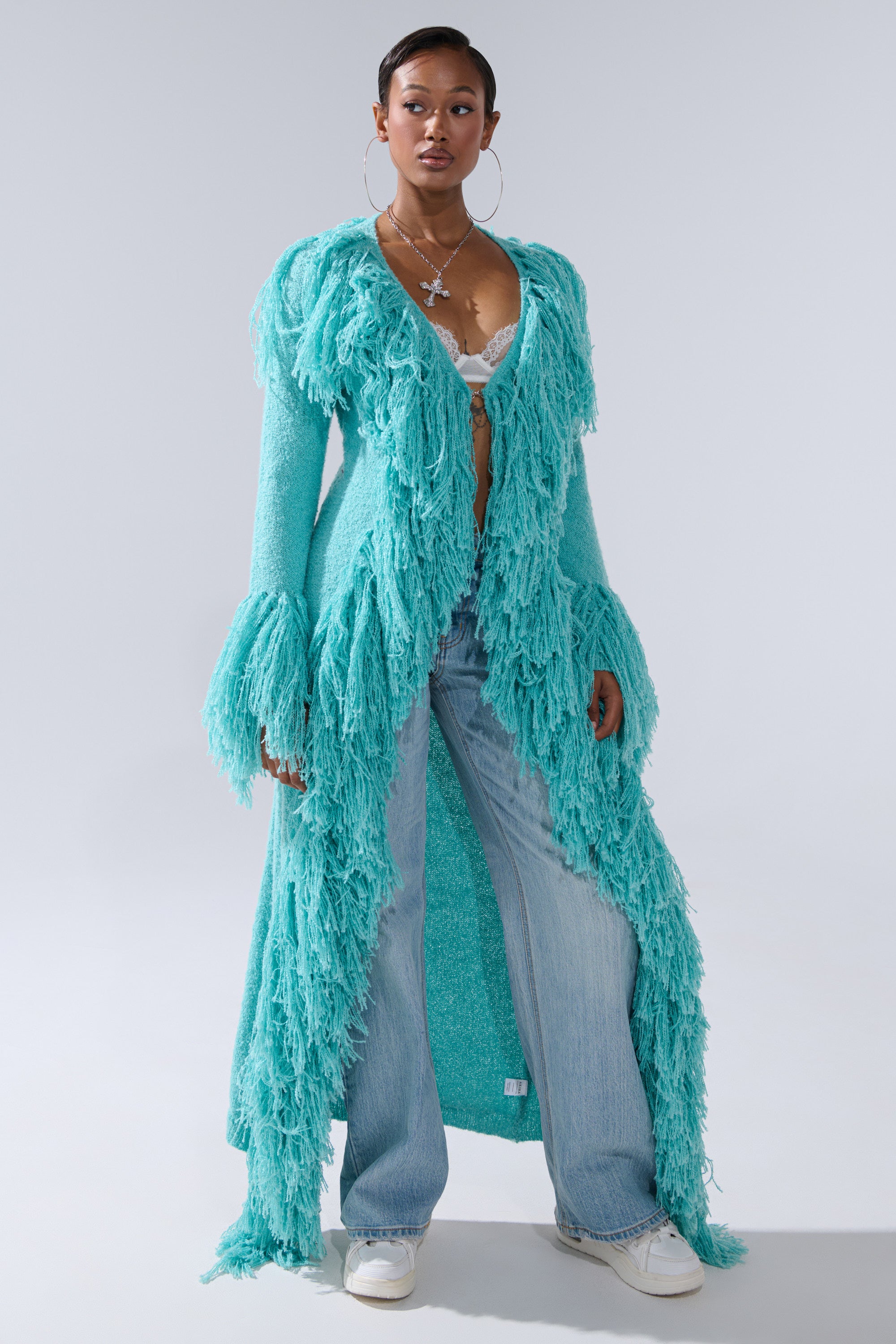 A woman stands confidently against a gray backdrop, wearing the BURRR IT'S COLD OUTSIDE FRINGE OVERSIZED SWEATER IN TURQUOISE over a lace bralette, paired with wide-leg jeans and white sneakers.