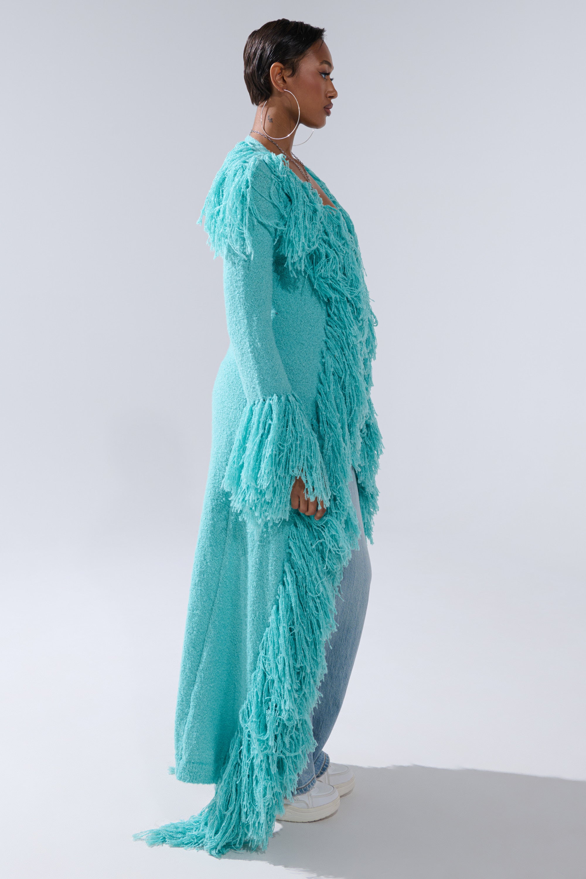 A person stands in profile wearing the BURRR IT'S COLD OUTSIDE FRINGE OVERSIZED SWEATER IN TURQUOISE, paired with blue jeans and white sneakers, set against a plain light background.
