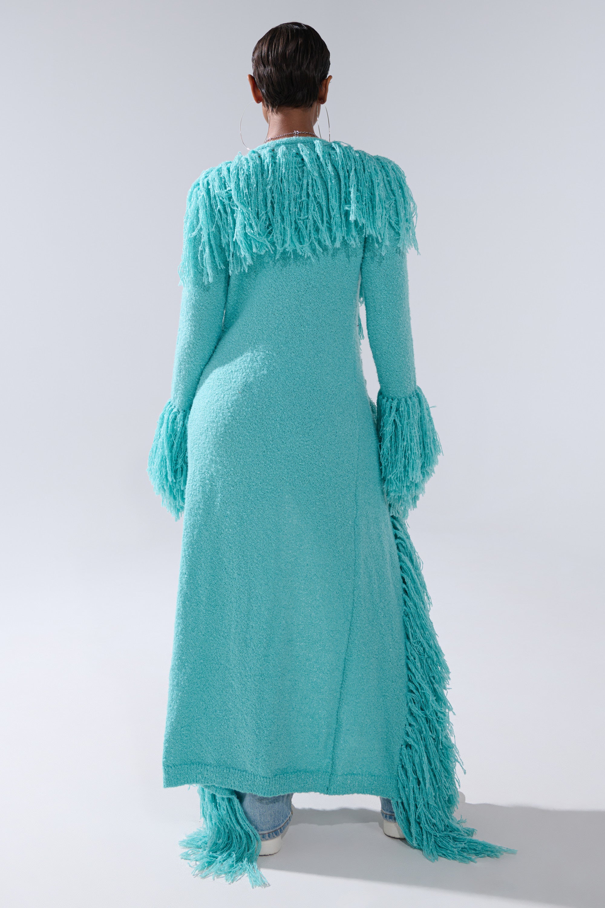 A person stands with their back to the camera, wearing the BURRR IT'S COLD OUTSIDE FRINGE OVERSIZED SWEATER IN TURQUOISE with fringe on the shoulders, cuffs, and hem, paired with matching shoes against a plain light gray background.
