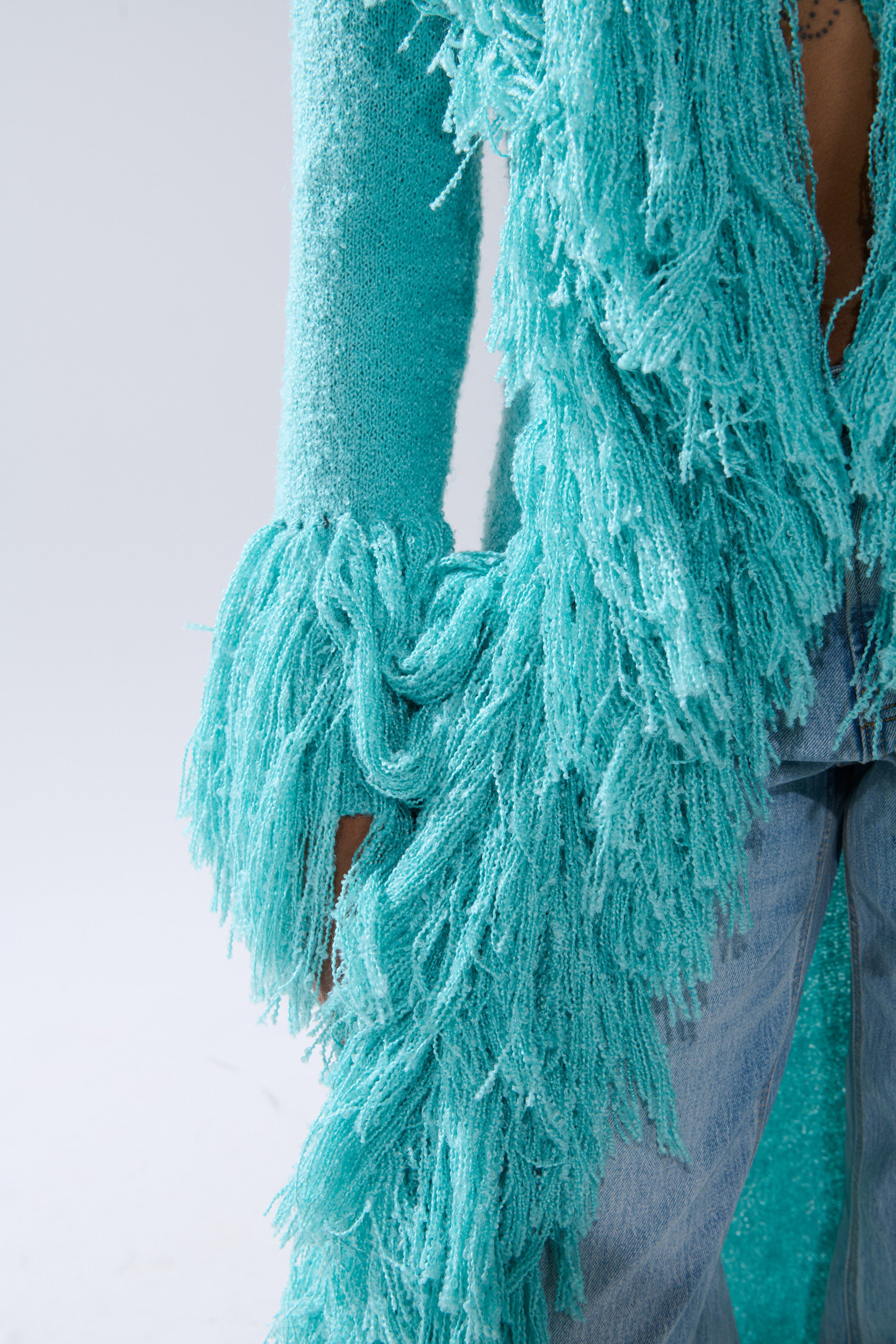 Someone wears light blue jeans and the BURRR IT'S COLD OUTSIDE FRINGE OVERSIZED SWEATER IN TURQUOISE, featuring long, shaggy fringe details, with their hand partially visible by their side.