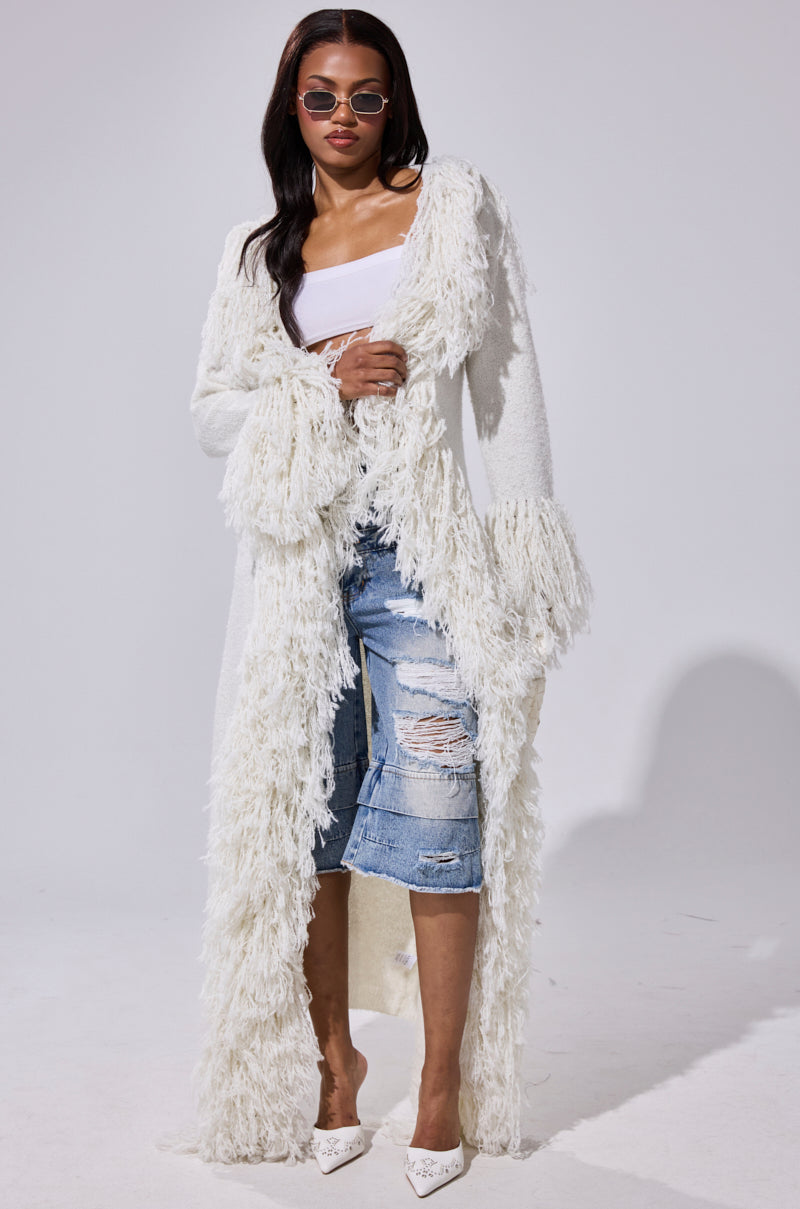 A woman poses confidently against a plain light background, styled in sunglasses, a white top, ripped denim jeans, white heels, and the BURRR IT'S COLD OUTSIDE FRINGE OVERSIZED SWEATER IN IVORY.