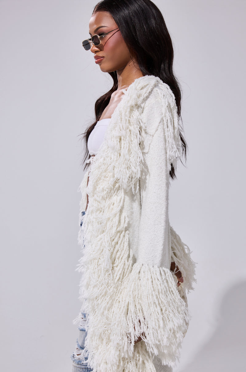 A woman in sunglasses models the BURRR IT'S COLD OUTSIDE FRINGE OVERSIZED SWEATER IN IVORY over a white crop top and light blue ripped jeans, standing sideways against a plain gray background.