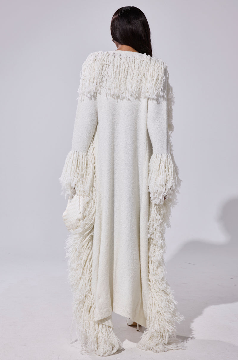 A person with long dark hair faces away, wearing the BURRR IT'S COLD OUTSIDE FRINGE OVERSIZED SWEATER IN IVORY featuring textured white fringe on the shoulders, sleeves, and hem, holding a small white bag against a plain background.