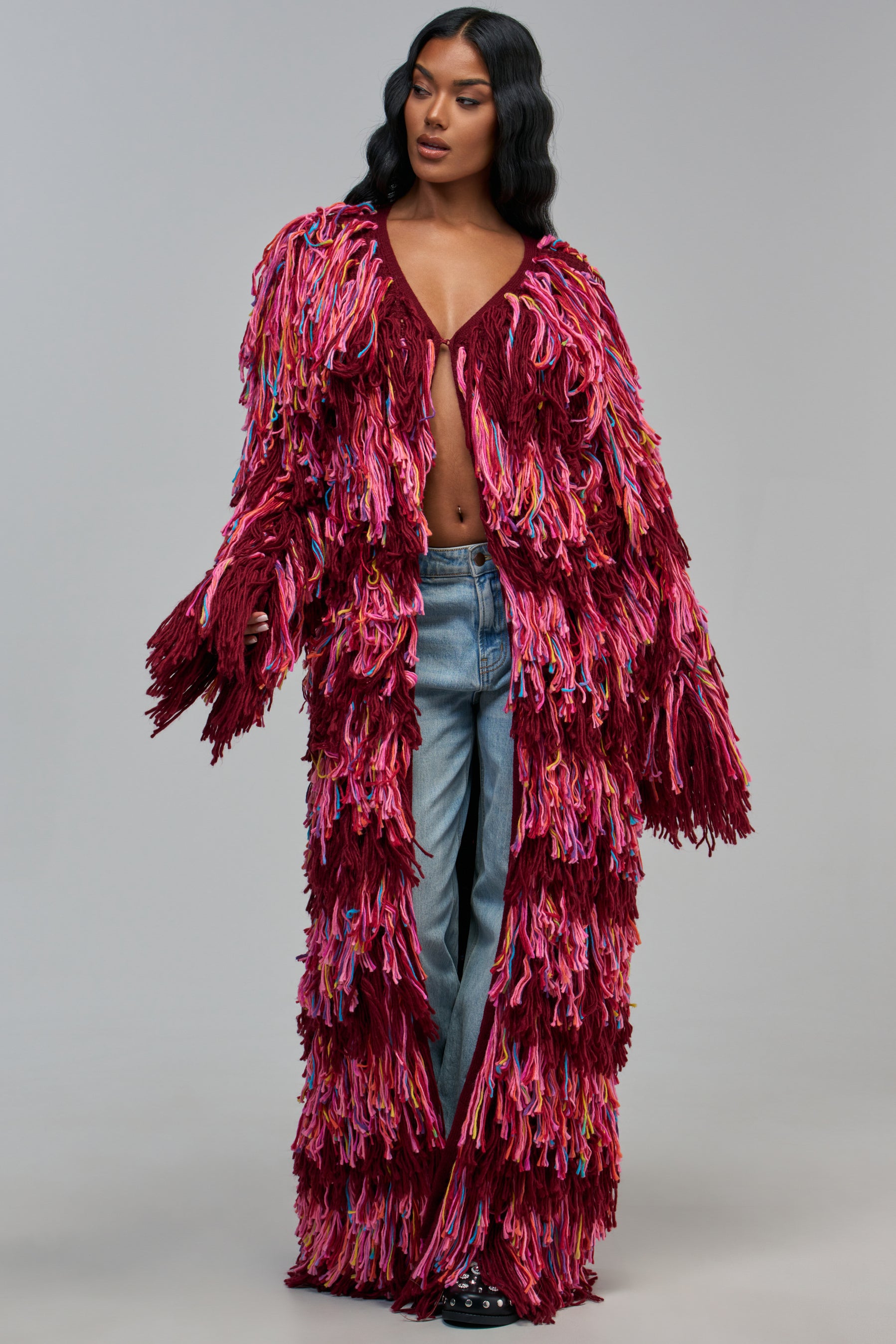 A person wears the SASHA LONG FRINGE CARDIGAN IN BURGUNDY, a floor-length style covered in multicolored yarn fringe, paired with blue jeans for a stylish fall look.
