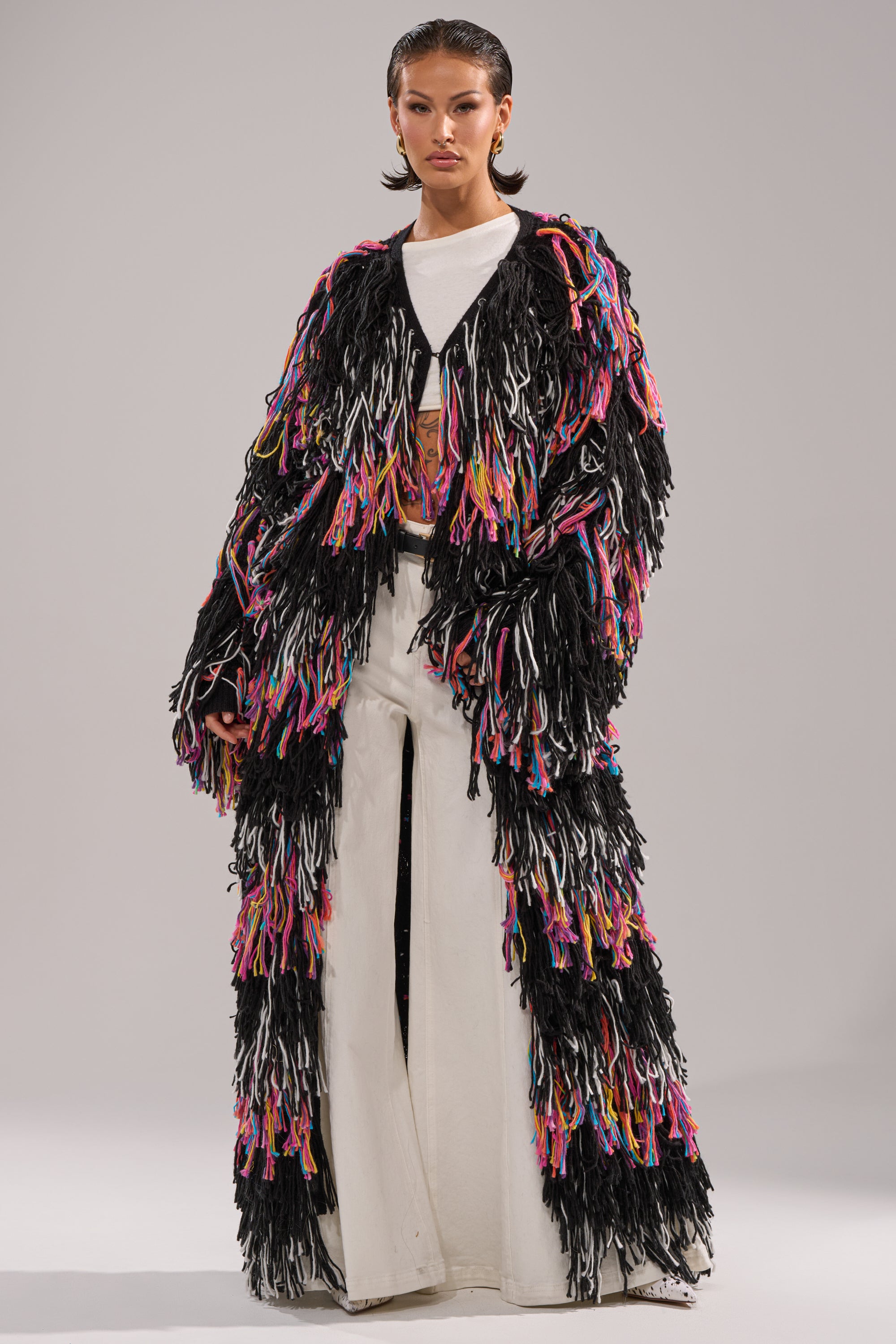 A person wears the SASHA LONG FRINGE CARDIGAN, featuring multicolored yarn fringe, over a white outfit with wide-leg pants, posing against a plain gray background.