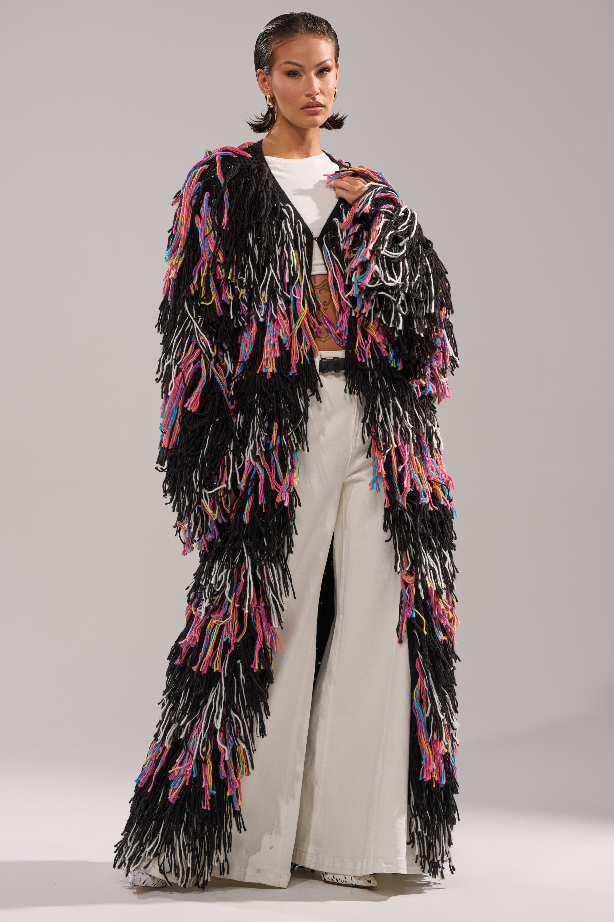 A person poses confidently against a light gray background wearing the SASHA LONG FRINGE CARDIGAN, a floor-length sweater featuring colorful yarn accents and long fringe, styled over a white top and wide-legged white pants.
