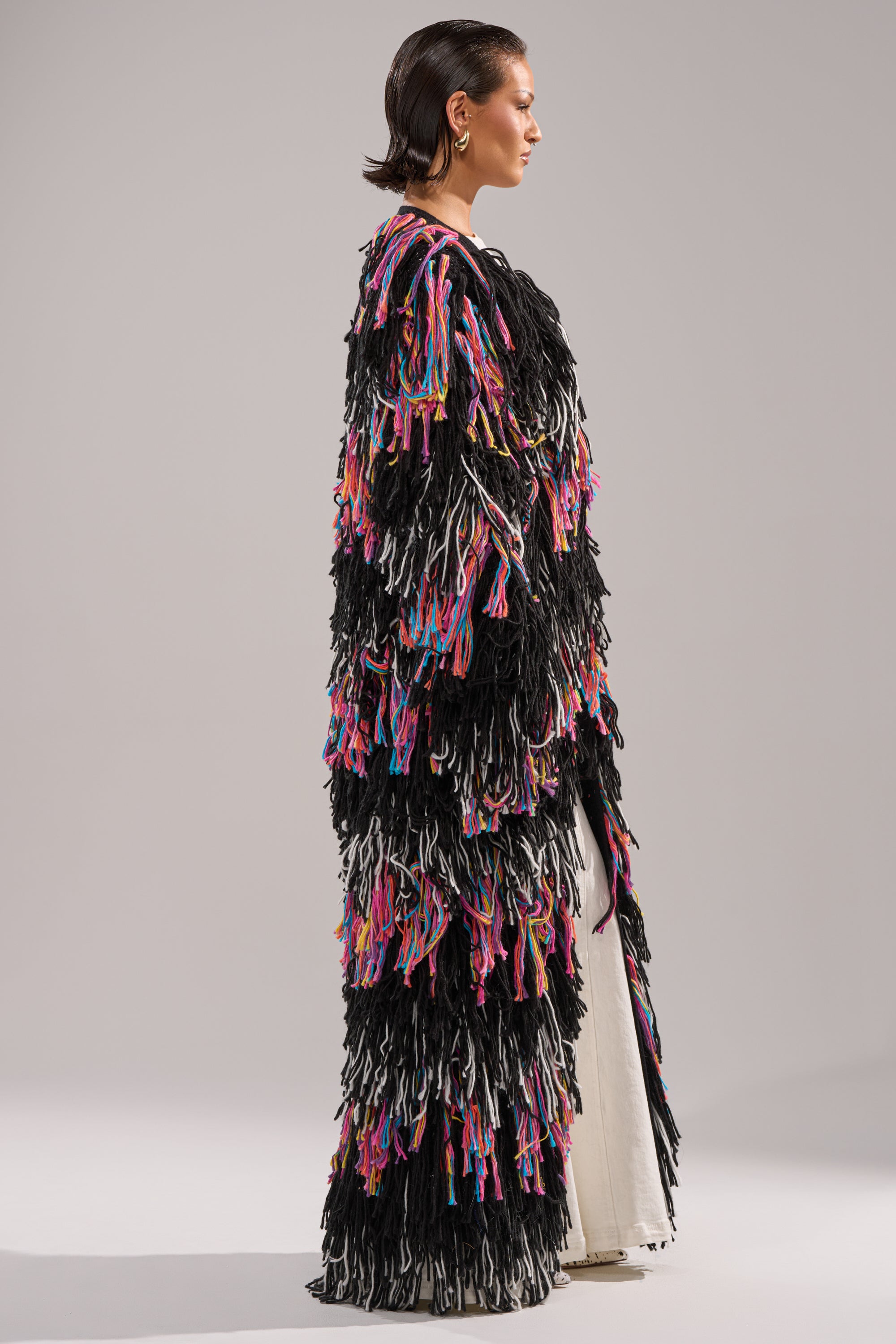 A person stands in profile wearing the SASHA LONG FRINGE CARDIGAN, a black, floor-length sweater adorned with colorful layered tassels, over a white outfit against a plain gray background.