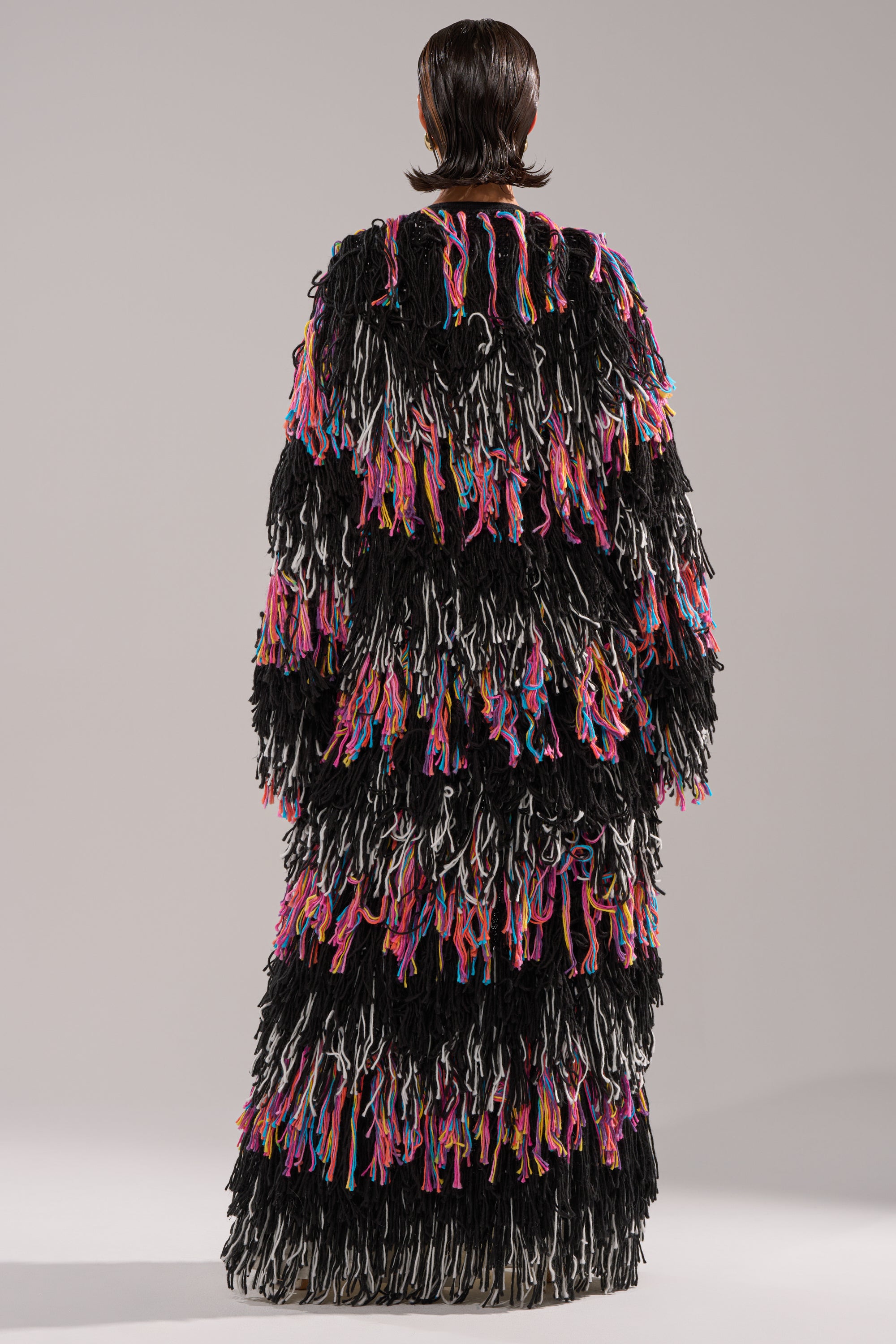 A person with short, slicked-back hair faces away, wearing the SASHA LONG FRINGE CARDIGAN—a floor-length sweater crafted from thick, multicolored yarn fringes in black, white, pink, orange, and blue for a textured, layered look.