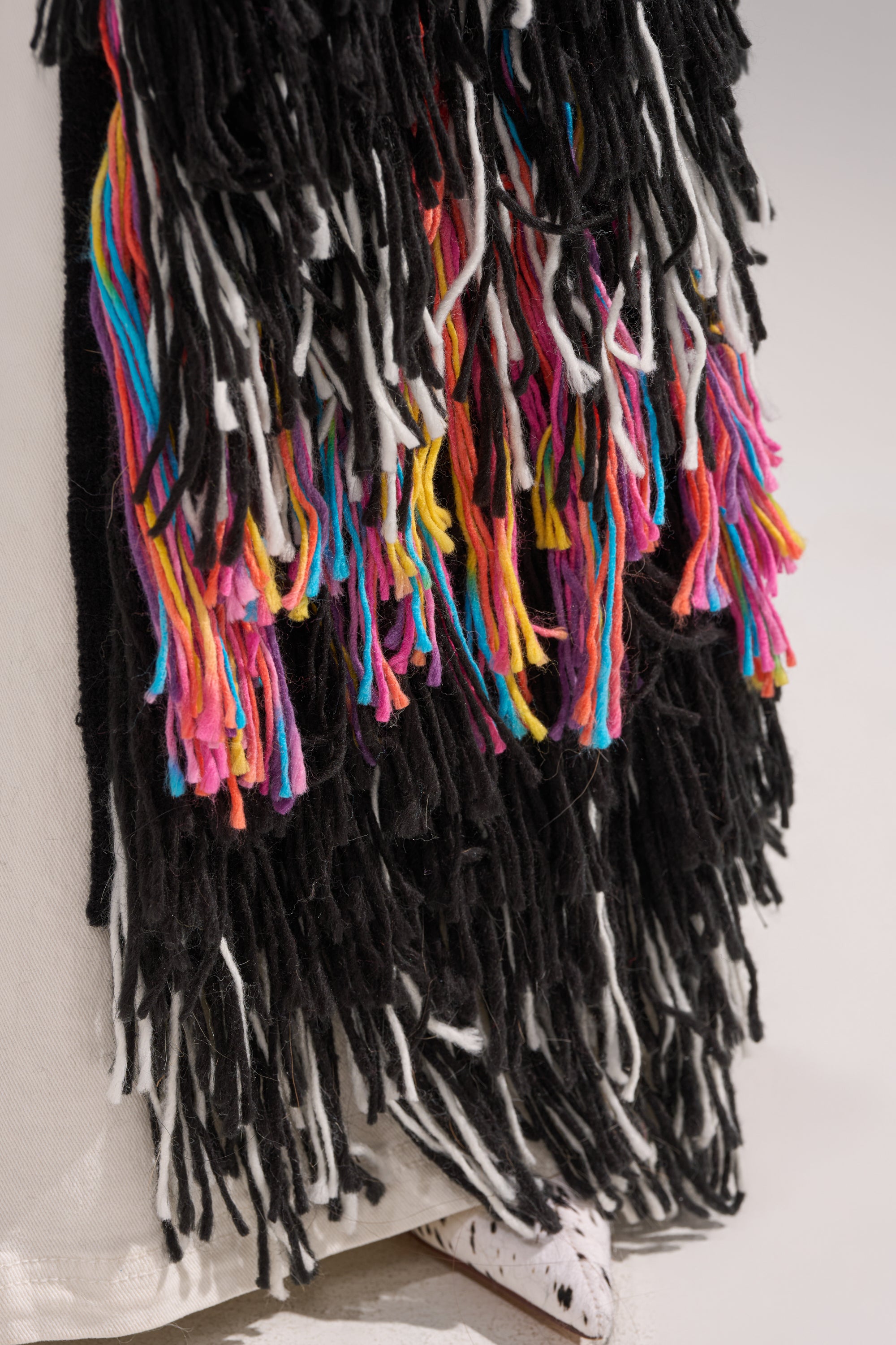 Close-up of the SASHA LONG FRINGE CARDIGAN, featuring thick tasseled yarn in black, white, pink, yellow, and blue over a white background. The multicolored fringe strands create a textured and layered look.