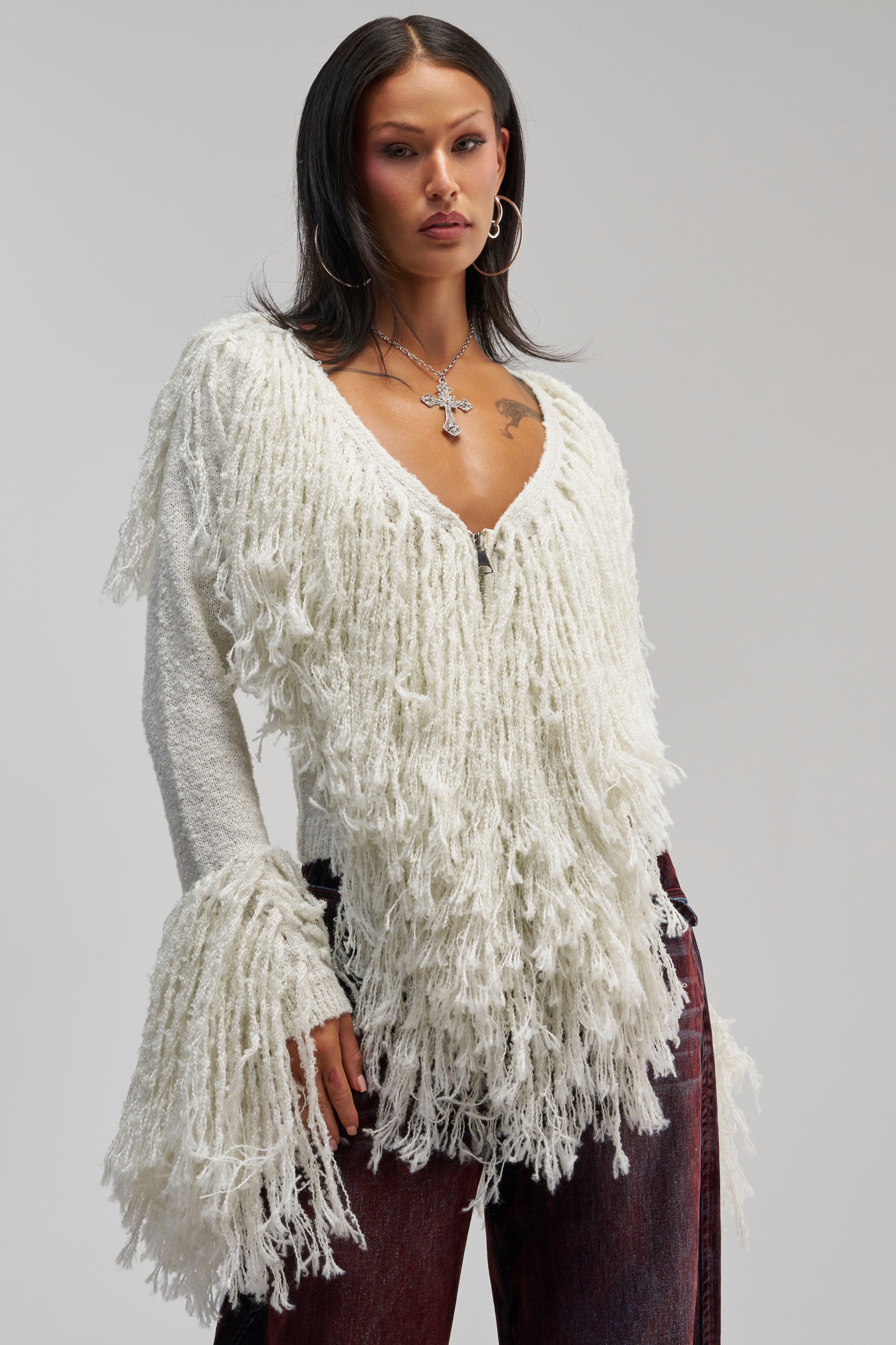 A woman wearing the AT YOUR BEST FRINGE CARDIGAN—a textured, off-white statement piece with fringed details and flared sleeves—pairs it with dark red pants as she stands against a plain background. With her long dark hair, hoop earrings, and bold cross necklace, she perfectly captures effortless winter styling.