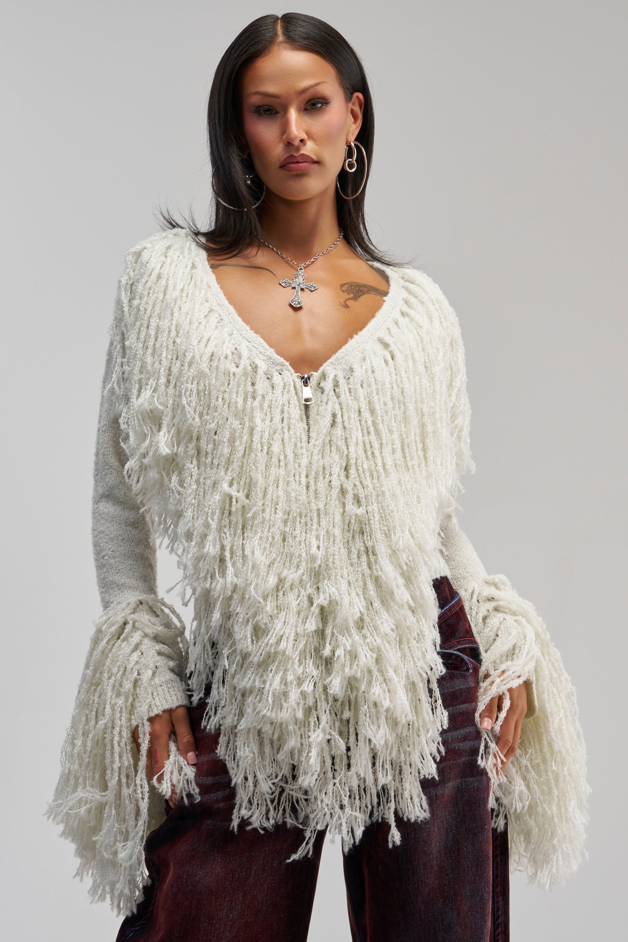 Wearing the AT YOUR BEST FRINGE CARDIGAN—a textured, off-white statement sweater—over dark red velvet pants, this woman accessorizes with hoop earrings and a cross necklace, standing against a plain background for chic winter styling.