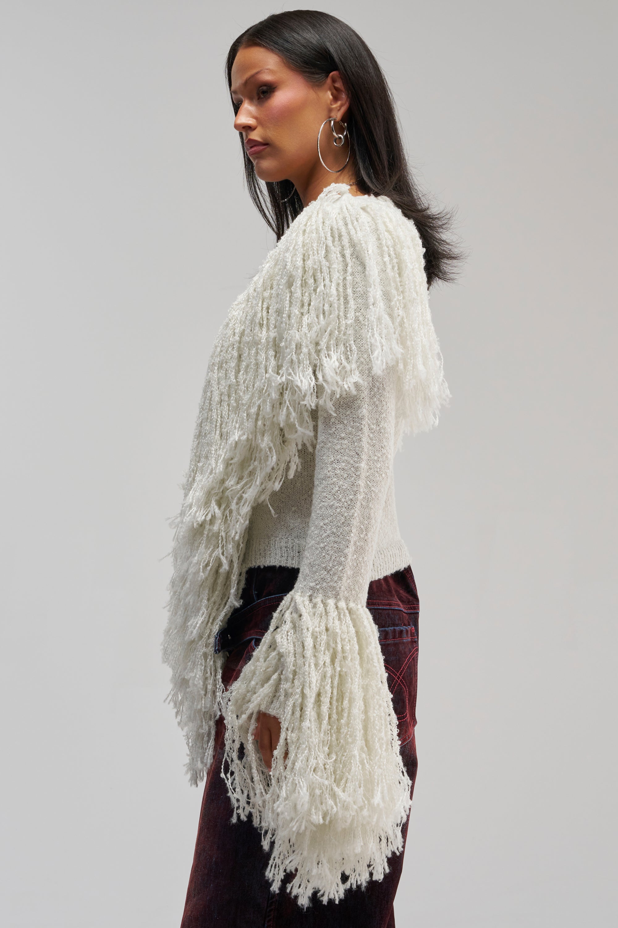 A woman in profile wears the AT YOUR BEST FRINGE CARDIGAN, a textured statement piece featuring long white fringe details on the sleeves and front, paired with dark pants. She accessorizes with hoop earrings and has straight, shoulder-length dark hair—perfect for chic winter styling.