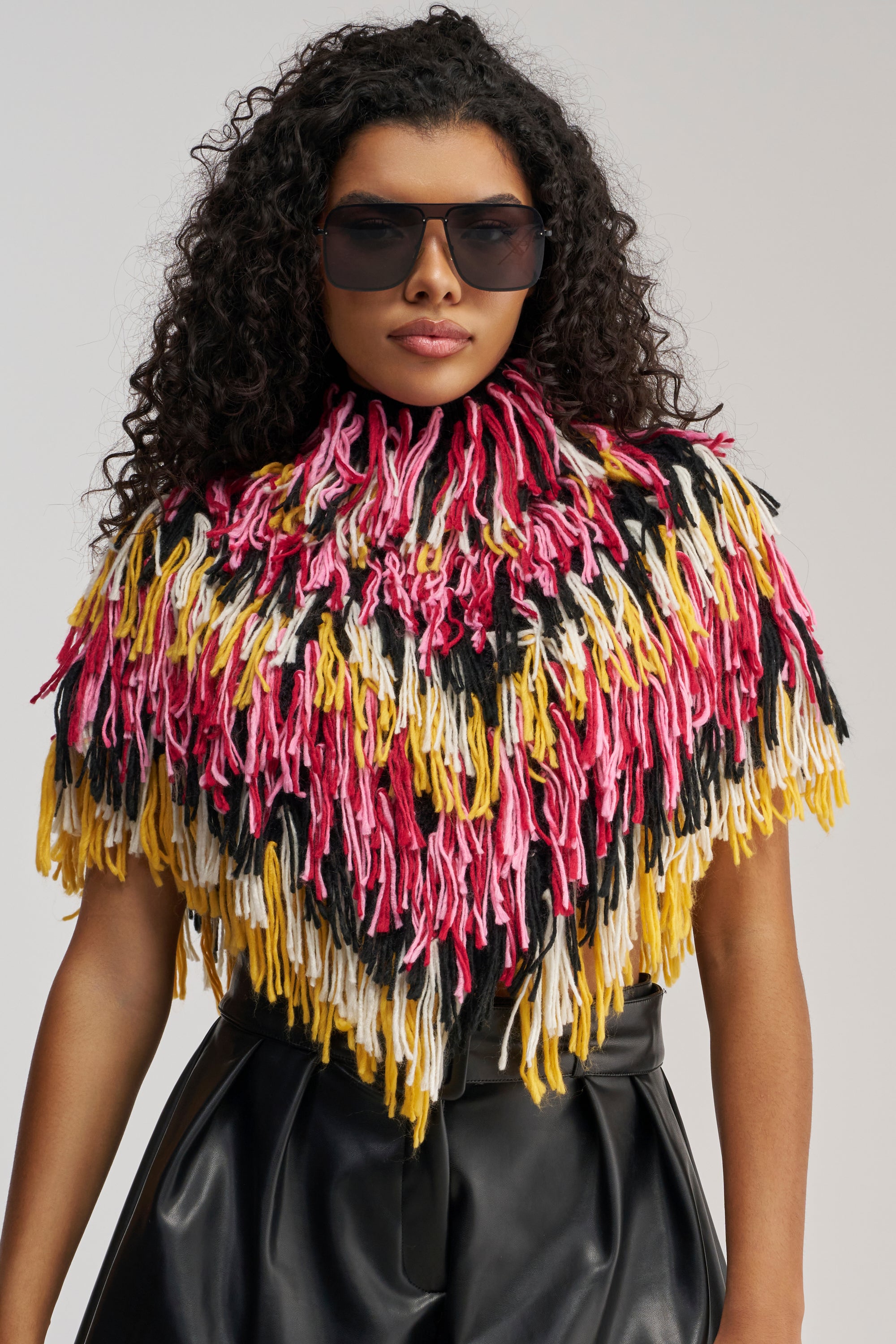 A person with curly hair wears dark sunglasses and the WILD SIDE CROPPED FRINGE PONCHO, which features multicolored fringe in pink, yellow, black, and white, paired with high-waisted black pants against a plain background.