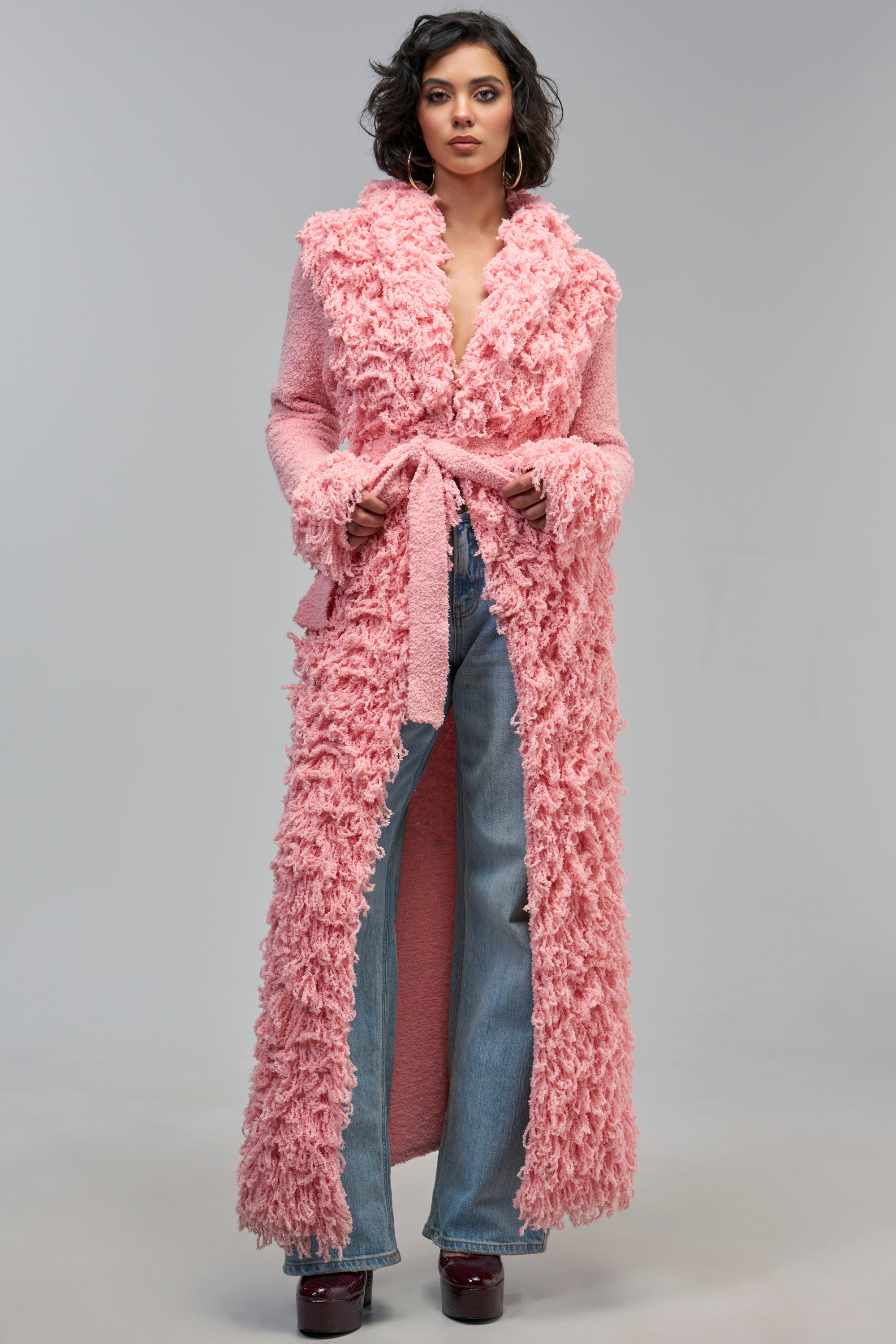 A model wears a light pink shaggy fringe COMFY COZY KNIT CARDIGAN with a tie waist over blue jeans.