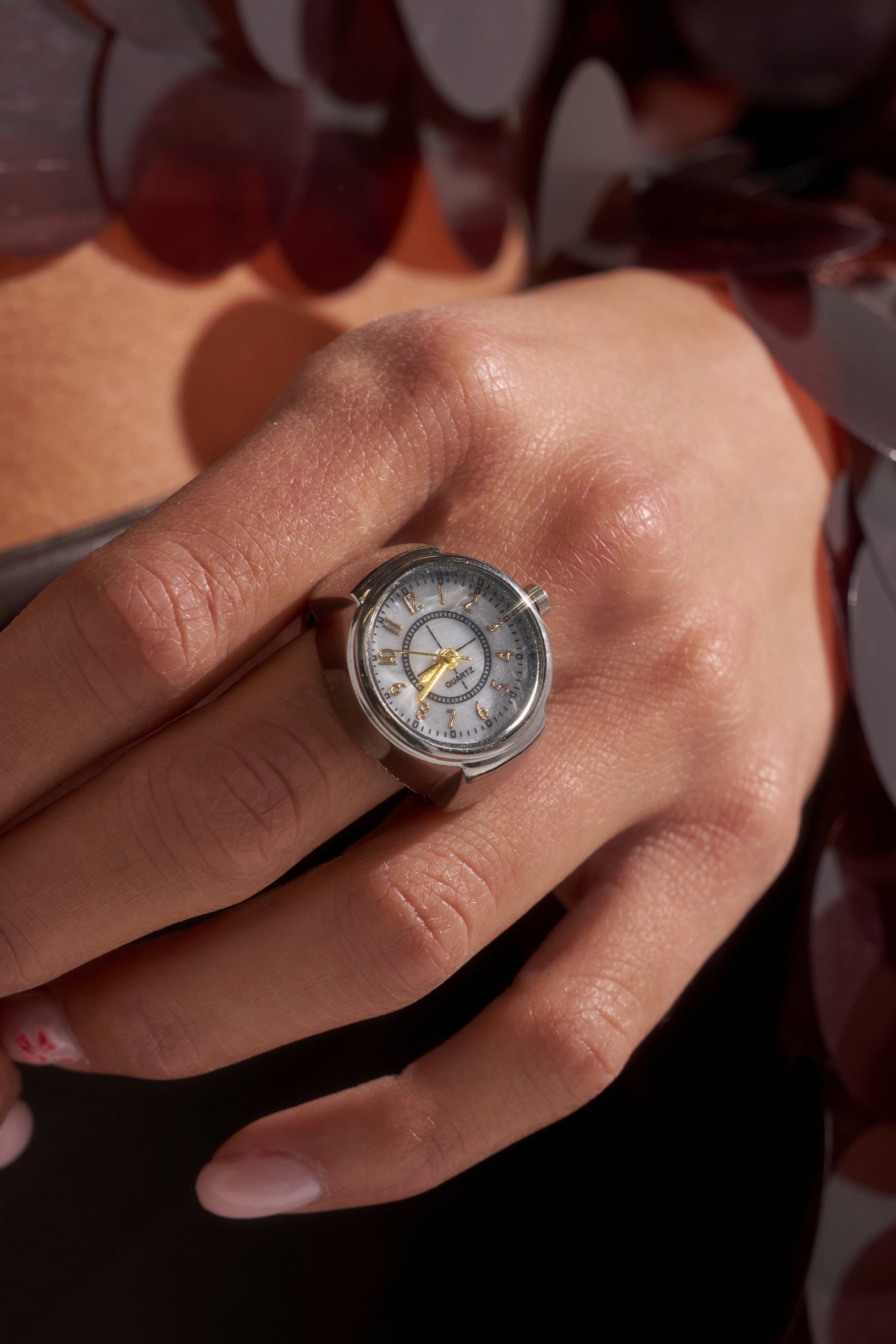 A close-up of a hand wearing the ITS TIME RING, a unique silver accessory crafted to resemble a small analog clock with visible hands and numbers, set against a blurred background. 4